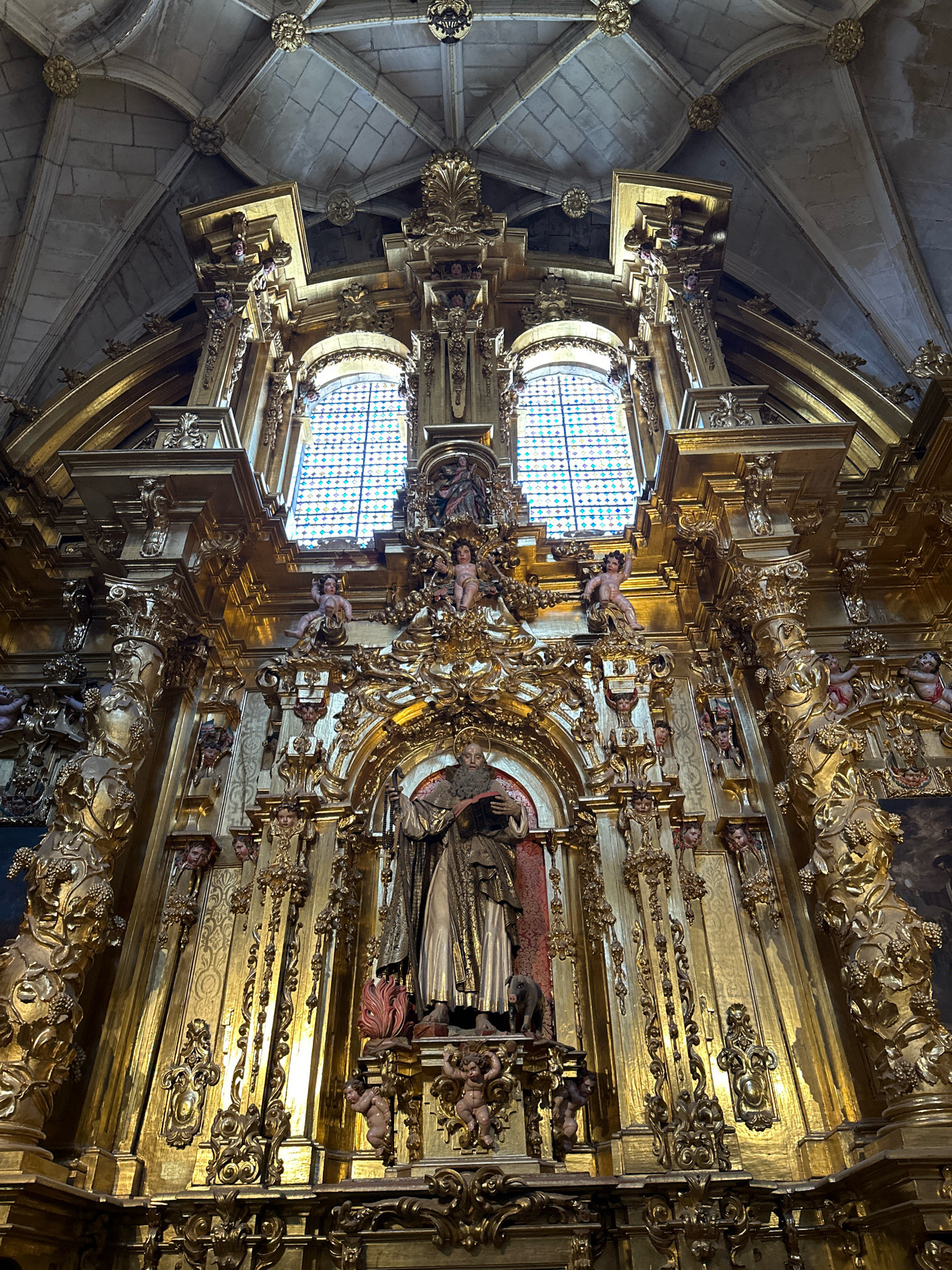 The image depicts the ornate golden altarpiece of a church, likely within the city of La Lastrilla, Spain. The altarpiece is a monumental structure of carved and gilded wood, reaching towards the stone vaulted ceiling. At its center, a statue of a bearded male figure, likely a saint, is prominently displayed. This figure wears a long robe and holds a book. To the left of the saint, a dark-colored animal, possibly a pig, is depicted. Surrounding the central figure and extending upwards are numerous decorative elements, including cherubic figures, intricate scrollwork, and architectural moldings, all rendered in glistening gold. Two large windows with stained glass are visible behind the upper portion of the altarpiece, casting colored light onto the golden surface. The overall impression is one of opulence and religious grandeur.