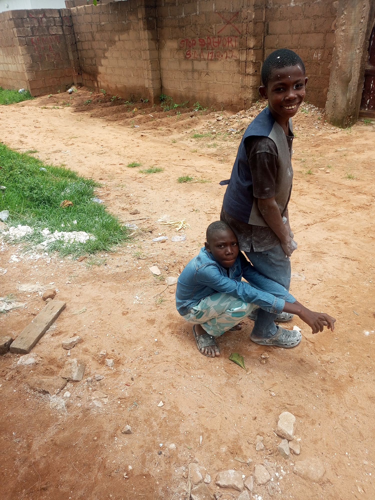 Two young boys are standing and crouching in a dirt-filled yard with a brick wall in the background. The boy standing is smiling and looking at the camera, while the boy crouching is looking down at the ground. Both boys are wearing casual clothing, and the ground around them is littered with debris. The brick wall has graffiti on it. The location is described as Tempure, Nigeria.