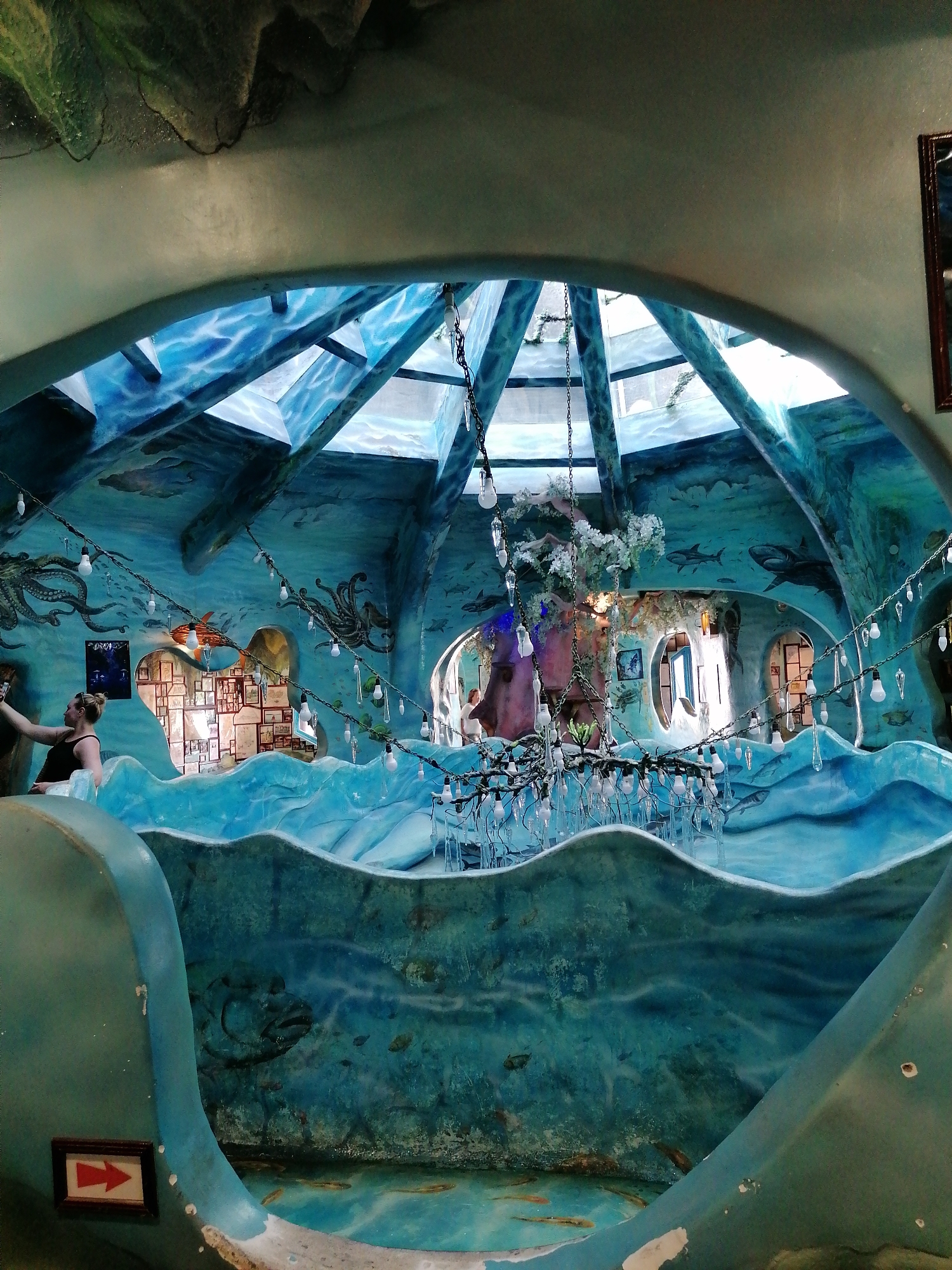 The image shows the interior of the Crazy House, also known as Hang Nga guesthouse, in Da Lat, Vietnam. The architecture is whimsical and surreal, resembling organic forms like trees and caves. The scene is predominantly turquoise and blue, designed to simulate an underwater environment. The roof is constructed with a glass dome that allows natural light to filter in, illuminating painted depictions of sea creatures like sharks and octopuses on the walls. A woman is visible taking a picture with her phone. A chandelier with dangling light bulbs and icicle-like decorations hangs in the center of the room. Other smaller rooms can be seen through archways. A directional arrow sign is visible on the bottom left.