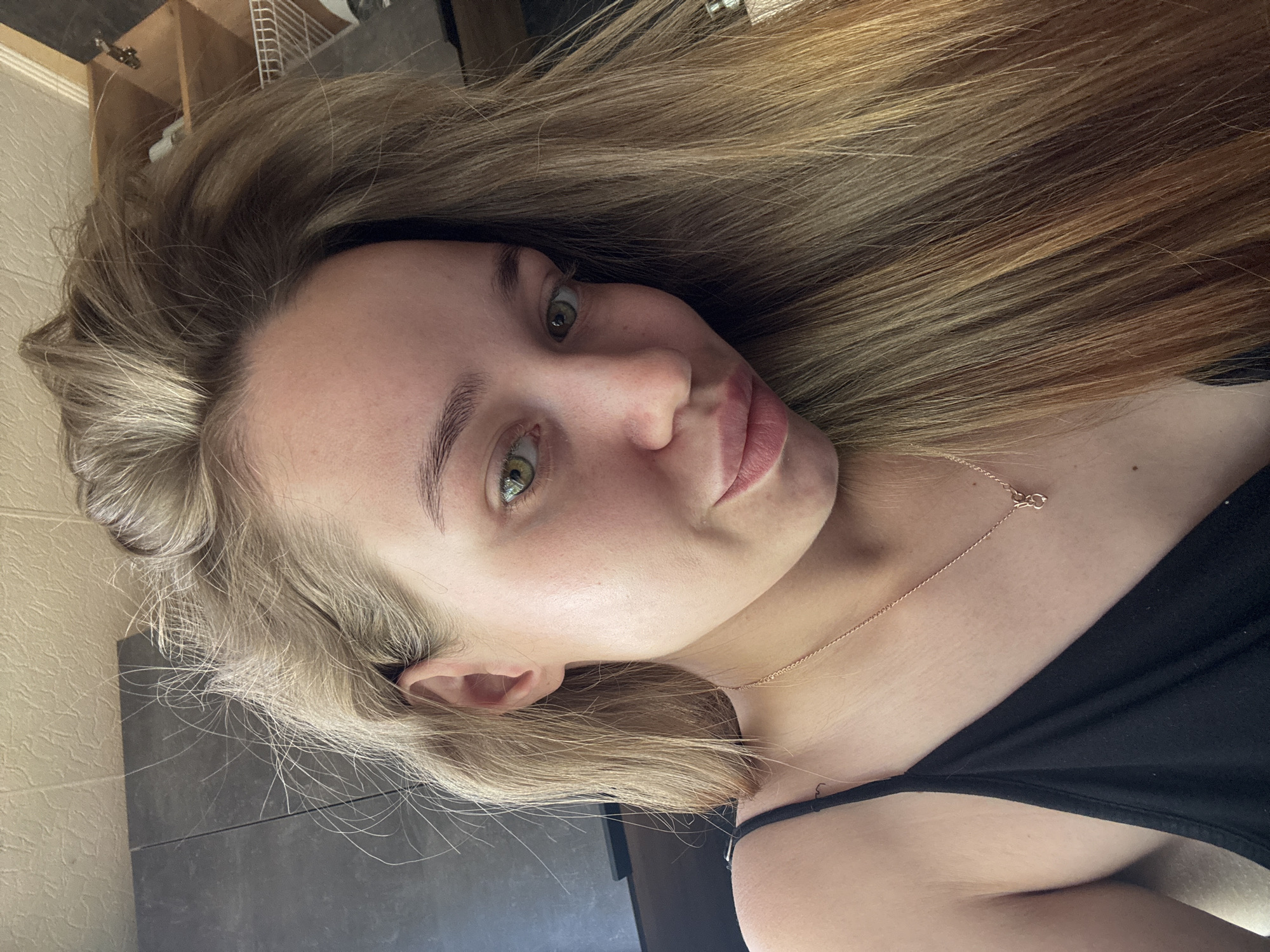 This is an angled, close-up portrait of a young woman, likely a selfie. Her head is tilted, with her long, light brown and blonde hair fanned out around her. She has fair skin, prominent green eyes, and a neutral expression with a slight pout. Her eyebrows are light and well-defined. She is wearing a simple black top or strap, and a delicate gold-colored chain necklace with a small, unidentifiable pendant rests on her chest.
The background indicates an indoor setting. To the left, a light-colored, textured wall is visible. Behind her head and to the right, there's a darker, smooth surface, possibly a piece of furniture or a countertop. In the upper background, parts of a wooden shelf or cabinet and a white grid-like object are discernible. The lighting appears to be soft, natural indoor light. No text is visible in the image.