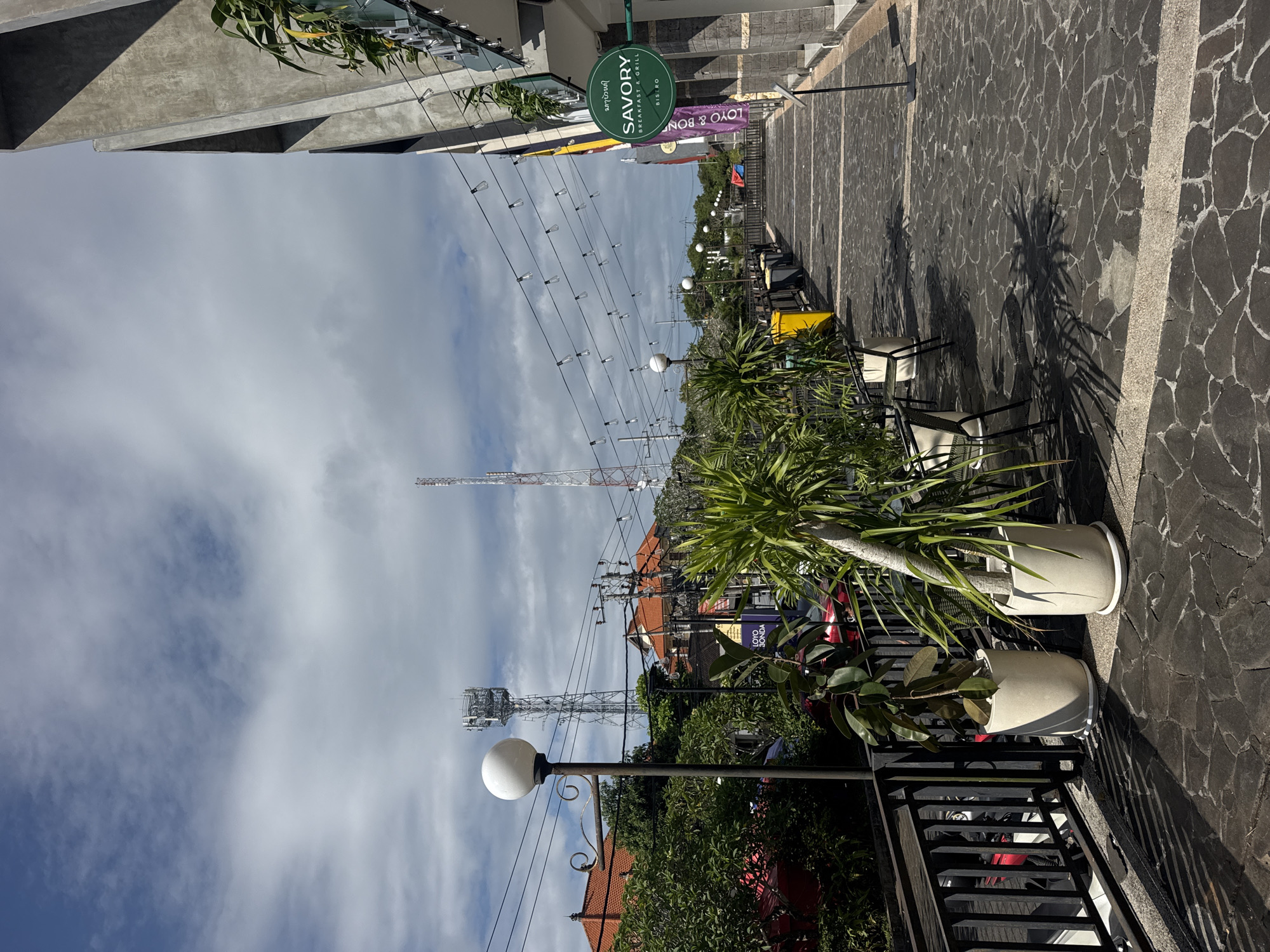 The image depicts a street scene in Kuta Selatan, Indonesia, likely a commercial or tourist area. The foreground features potted plants and a black metal railing, with a paved walkway beyond. A row of chairs and tables, likely belonging to a restaurant or cafe, lines the walkway. Overhead, string lights are suspended. Further down the street, signage is visible, including one for "SAVORY Breakfast & Grill". Utility poles and telecommunication towers are prominent in the background, set against a partly cloudy sky. Buildings with red tiled roofs are also visible. The overall impression is a blend of commercial activity and local infrastructure.