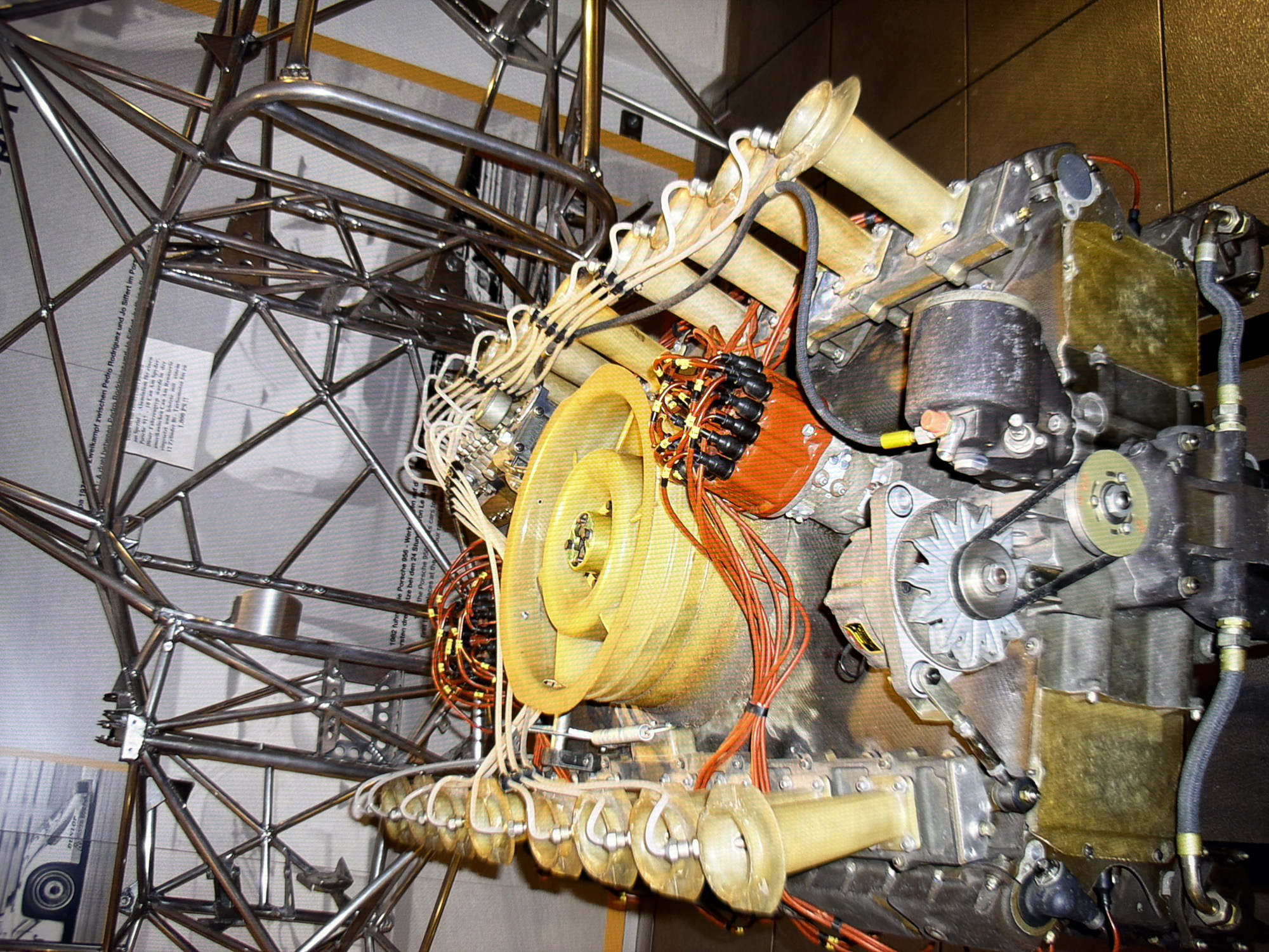 The image shows a close-up of a complex mechanical assembly, likely an engine or a part of a racing vehicle, with a prominent chrome-plated tubular frame structure behind it. The mechanical assembly features numerous wires, hoses, yellow cylindrical components, and an orange distributor cap with black spark plug boots. The frame is made of interconnected metal tubes, suggesting a lightweight and strong construction. There is a printed sign partially visible in the background with text in German. The lighting appears to be artificial, casting shadows on the components. The floor beneath the assembly is tiled. There are no people visible in the image. The image quality is good, with clear details of the mechanical parts.