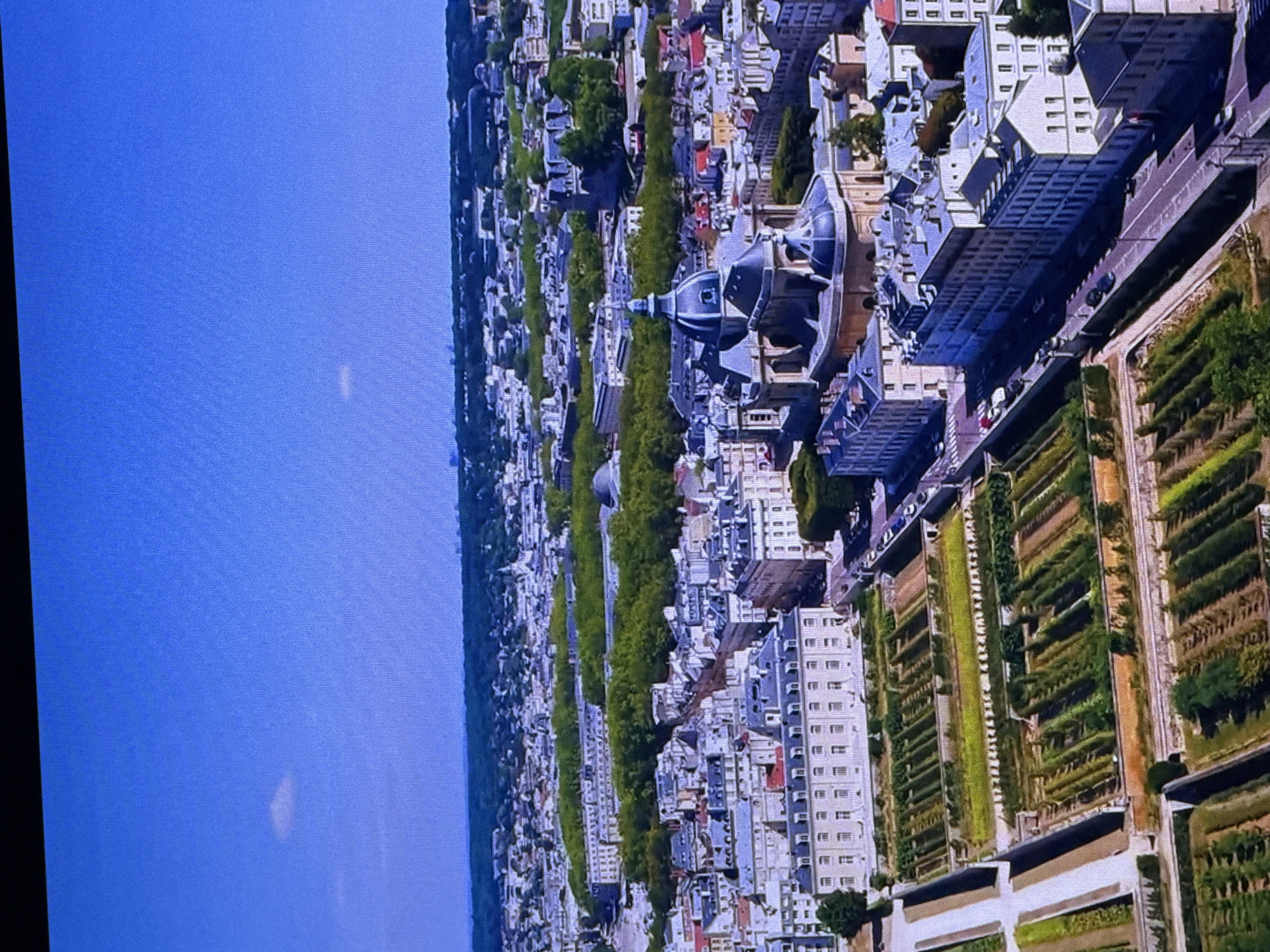 The image is an aerial view of a long, tree-lined avenue leading to a large building with a domed roof. The avenue is bordered by ornate buildings on one side and what appear to be formal gardens on the other. The gardens are laid out in a geometric pattern with manicured hedges and planted beds. Several cars are visible on the road and in parking areas. The sky is a clear, bright blue, suggesting a sunny day. The architecture of the buildings, with their mansard roofs and classical detailing, suggests a European city, possibly Paris. However, given the prompt context of Dubai, this would be an unusual sight for that location. The image is oriented vertically, with the blue sky on the left and the cityscape on the right. There is no visible text in the image.