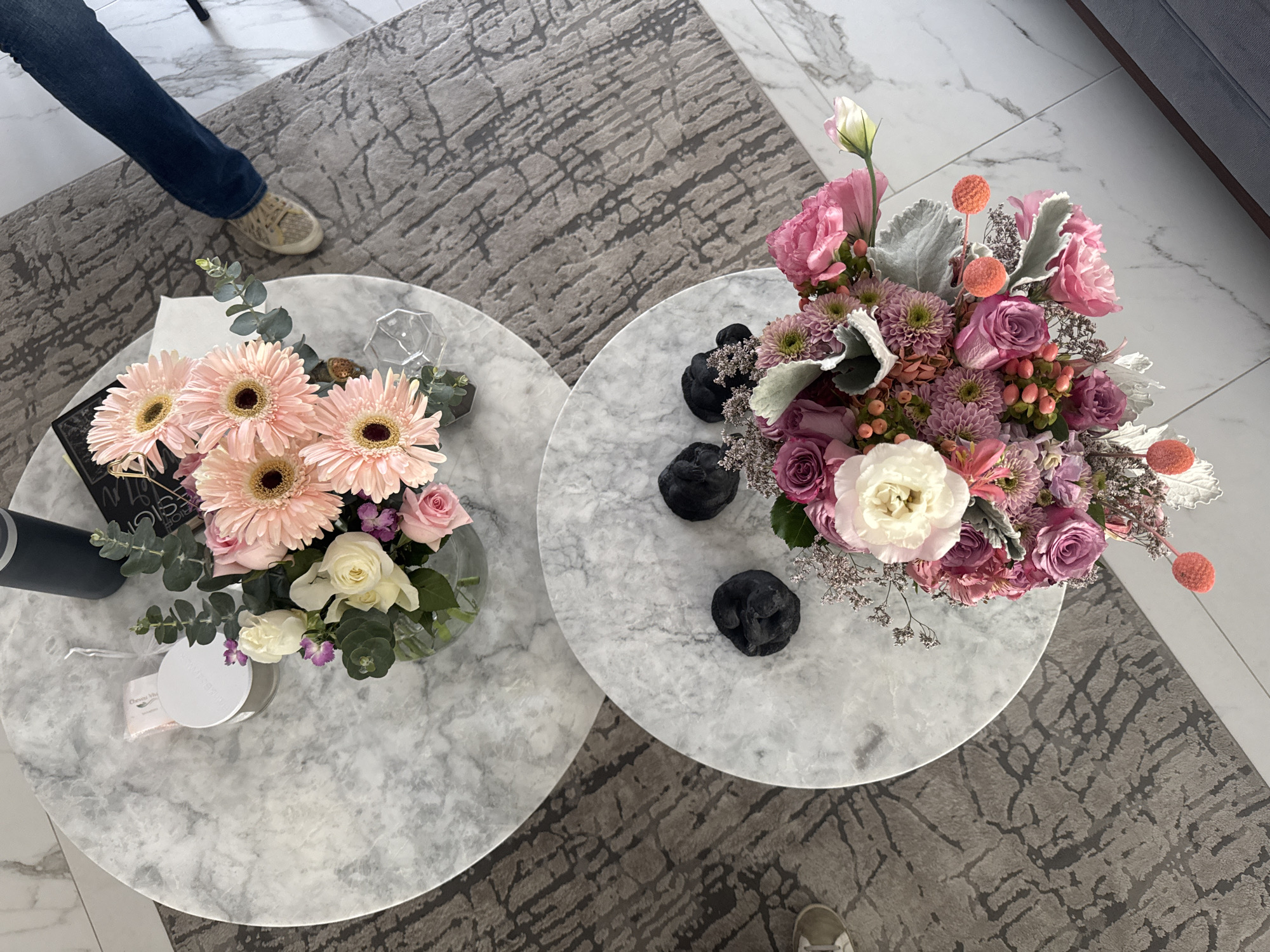 The image shows an overhead view of a room with two round marble-topped tables on a gray patterned rug. One table holds a bouquet of light pink gerbera daisies and white roses, a glass vase, a book with gold accents, a water bottle, a small, white, round container, and a crystal prism. The other table holds a bouquet of pink, purple, and white flowers, and four small black sculptures. Portions of two people are visible: one wearing jeans and sneakers, and another wearing sneakers. The room has white tile flooring near a gray couch. The location is Puebla, Mexico.