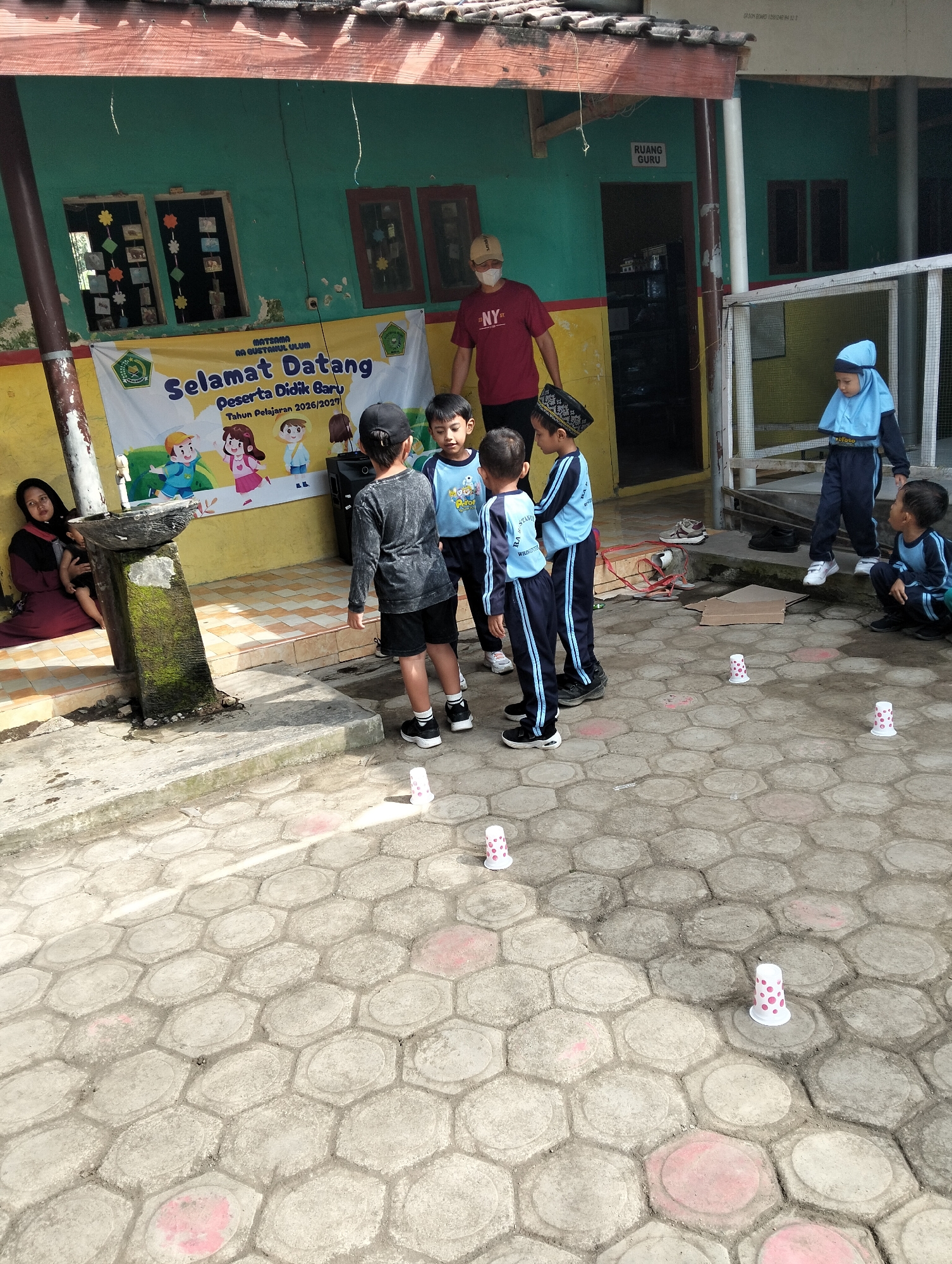 A group of young children, likely students, are gathered in an outdoor area in front of a building with green walls. Some of the children are wearing blue and white uniforms, while others are in casual attire. Several of the children appear to be interacting with each other or observing something on the ground. There are several white plastic cups with pink polka dots placed on the hexagonal paved ground, suggesting a game or activity is taking place. A banner with Indonesian text is visible on the wall of the building, indicating a welcome message for new students and the academic year. A woman in a maroon hijab and dress is seated on the ground to the left, holding a baby. A man in a red t-shirt and a mask is standing near a doorway on the right side of the image. The environment appears to be a school or community setting. The location is described as Desa Wringin Putih, Indonesia.