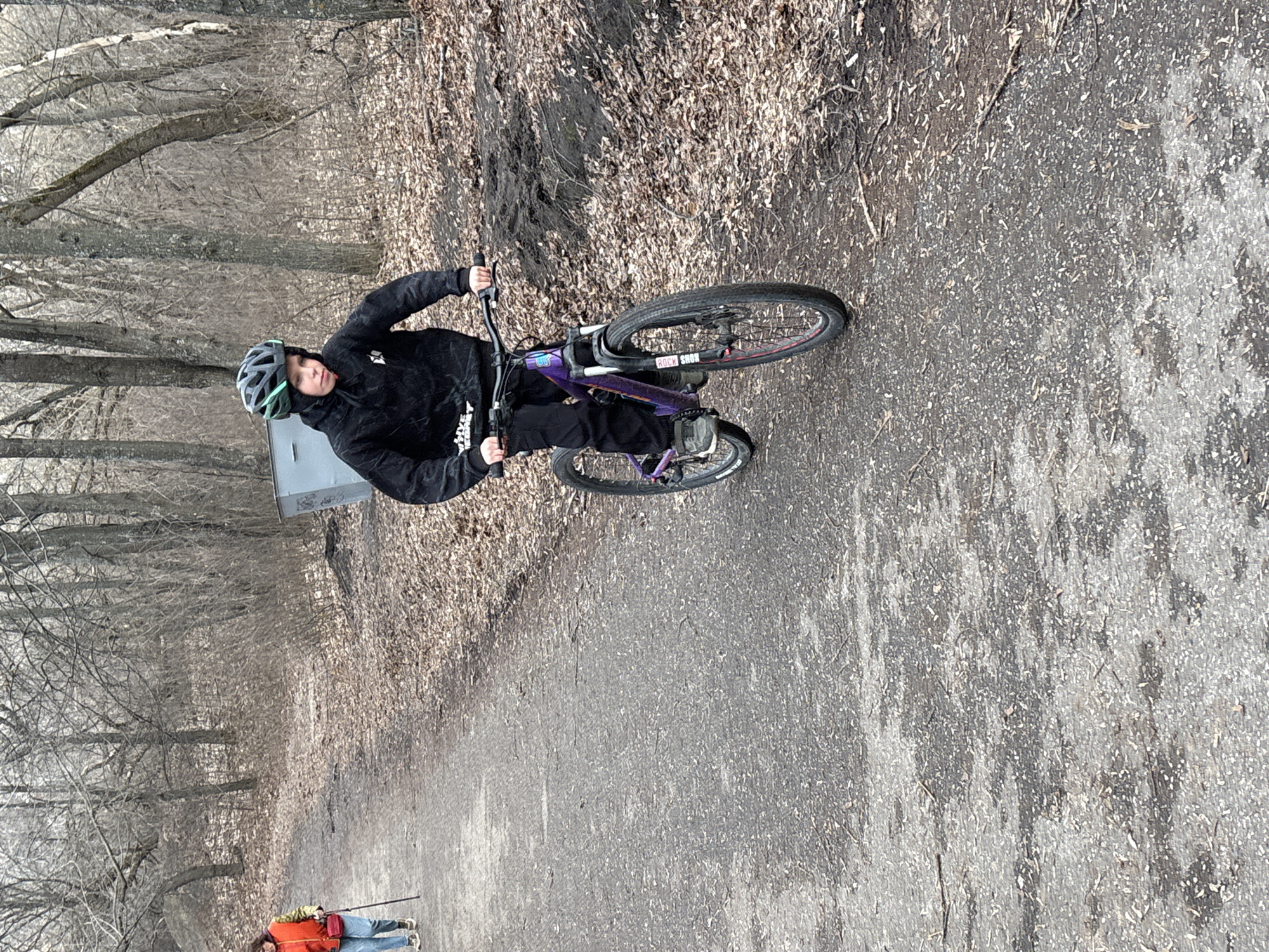 A person wearing a helmet and a black hoodie rides a purple and black bicycle on a paved path. The path is bordered by fallen leaves and a wooded area with bare trees. In the background, two people are walking. The location is likely a park or recreation area within the city of Ulyanovsk, Russia.