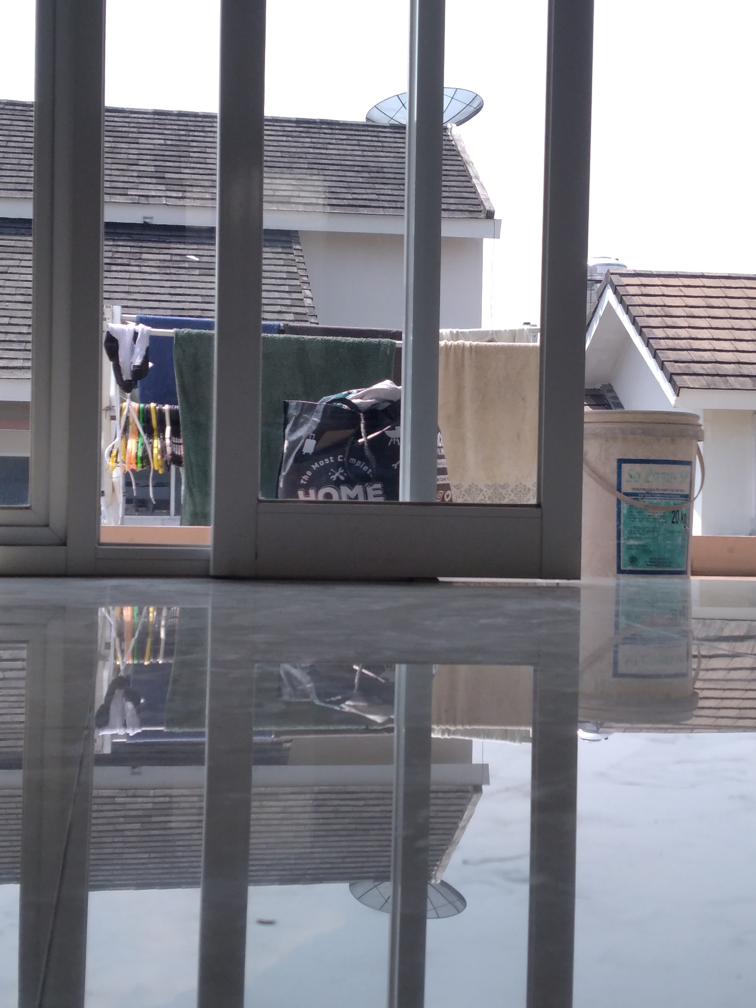 The image captures a view from inside a building, looking out through vertical white bars. In the foreground, a highly reflective, marble-like floor mirrors the scene above. Beyond the bars, clothes are hung out to dry on a clothesline, including socks, towels, and other garments. A black bag with the word "HOME" printed on it is visible among the laundry. In the background, two roofs of houses are visible, one with a satellite dish on top. To the right, a pale yellow bucket with a blue and green label is placed on a ledge. The sky is bright and overcast. This scene is likely located in Kaliwates, Indonesia, given the context of the surrounding environment and typical residential setups in the area.