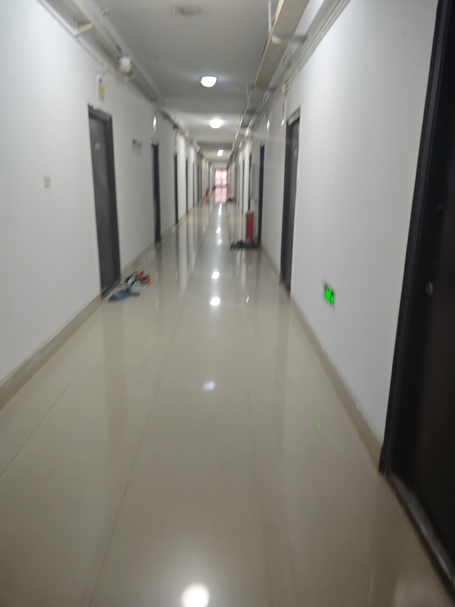 The image depicts a long, well-lit hallway with a tiled floor and white walls. Doors are visible on both sides of the hallway, leading to individual rooms. There are light fixtures along the ceiling. On the left side of the hallway, near one of the doors, a pair of blue sandals and a few other assorted footwear items are placed on the floor. Further down the hallway on the right wall, a small, green illuminated sign is affixed. The overall impression is that of a residential or dormitory hallway. The scene appears to be devoid of people.