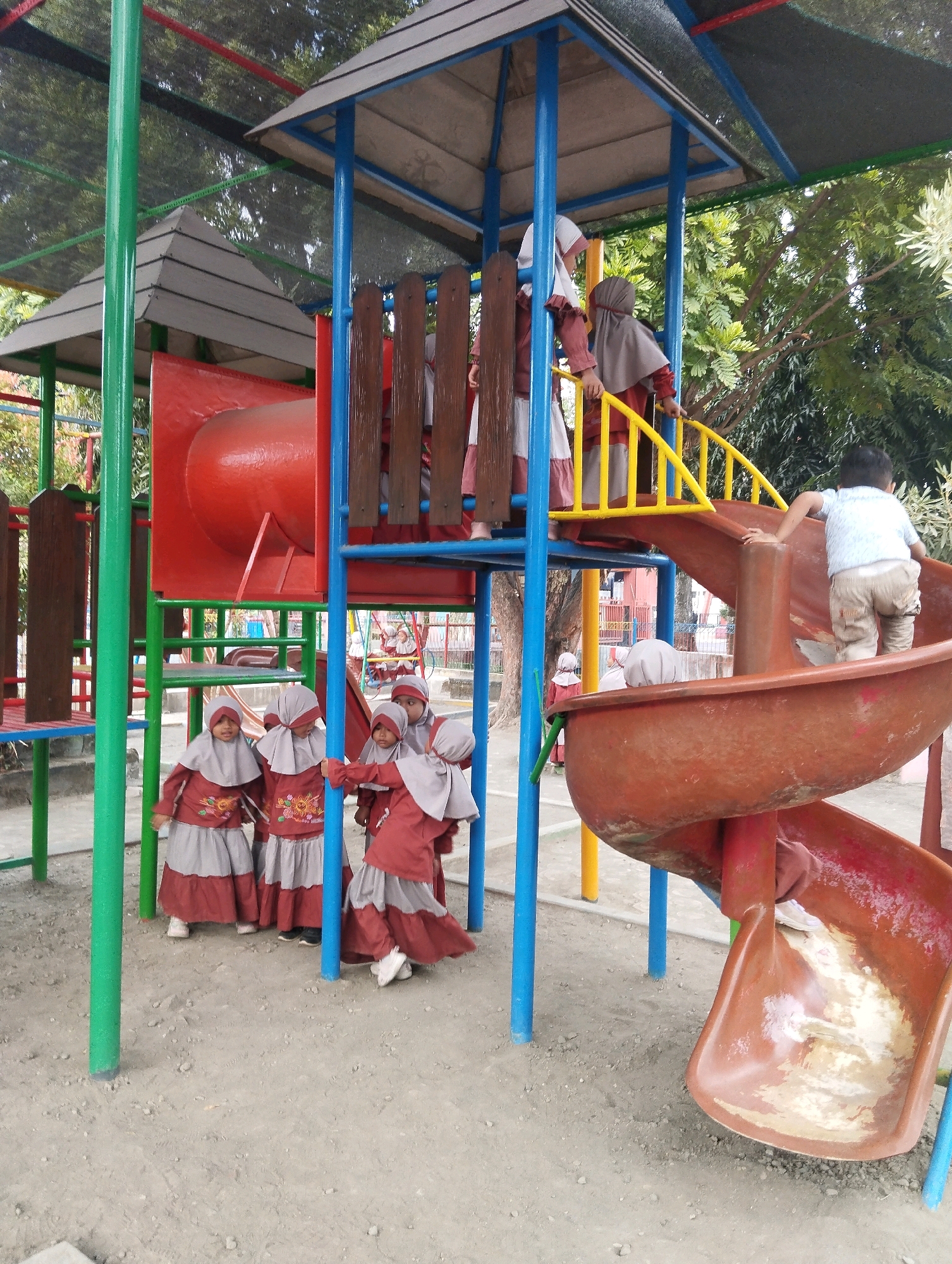 In the city of Blitar, Indonesia, a group of children are playing on a colorful, multi-level playground. The playground features a large red tubular slide, a yellow staircase, and various blue and green support poles. Several children, dressed in maroon and gray Islamic school uniforms with headscarves, are gathered at the bottom of the slide and on the lower platform. One child, wearing a light blue shirt and beige shorts, is in the process of sliding down the spiral tube. The ground is covered in sand and dirt. Trees and foliage are visible in the background, suggesting an outdoor setting, likely a schoolyard or park.