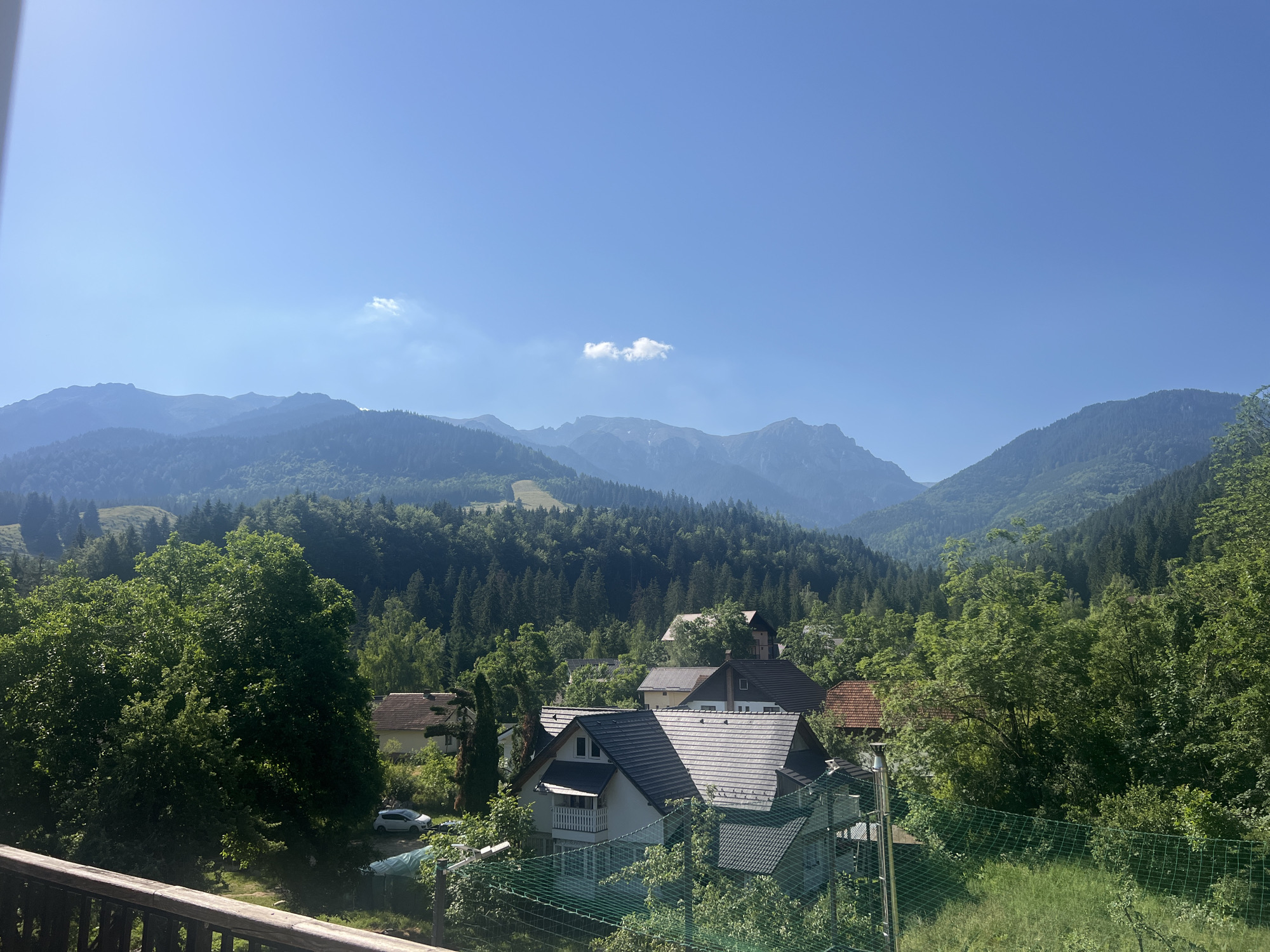 A wide-angle, eye-level shot captures a serene mountain village scene on a clear day. The foreground is dominated by a wooden railing, suggesting a balcony or deck. Beyond the railing, a verdant landscape unfolds, filled with lush trees and several houses with traditional pitched roofs. A white car is parked on a dirt road, partially obscured by foliage. The middle ground transitions into a dense forest that blankets rolling hills. In the background, majestic mountains rise, their peaks softened by a hazy blue sky dotted with a few small clouds. The overall impression is one of peaceful rural life nestled within a stunning natural setting.