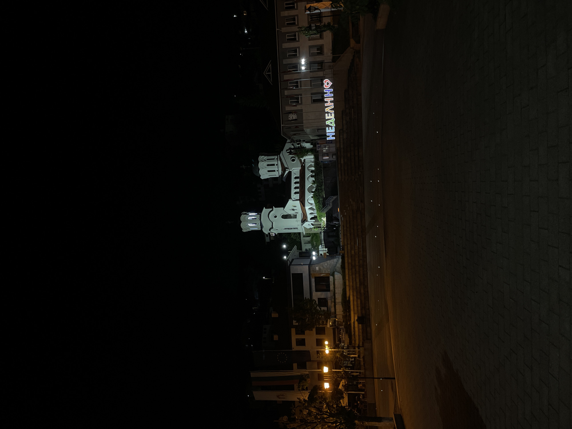 At night in Nedelino, Bulgaria, a white church with illuminated towers and arched walkways stands lit up. In the background, a building displays the city's name, "НЕДЕЛИНQ," in illuminated Cyrillic letters with a heart symbol. To the right, a dark paved area occupies a large portion of the image. Street lights illuminate other buildings and areas in the foreground, but the left side is mostly dark.