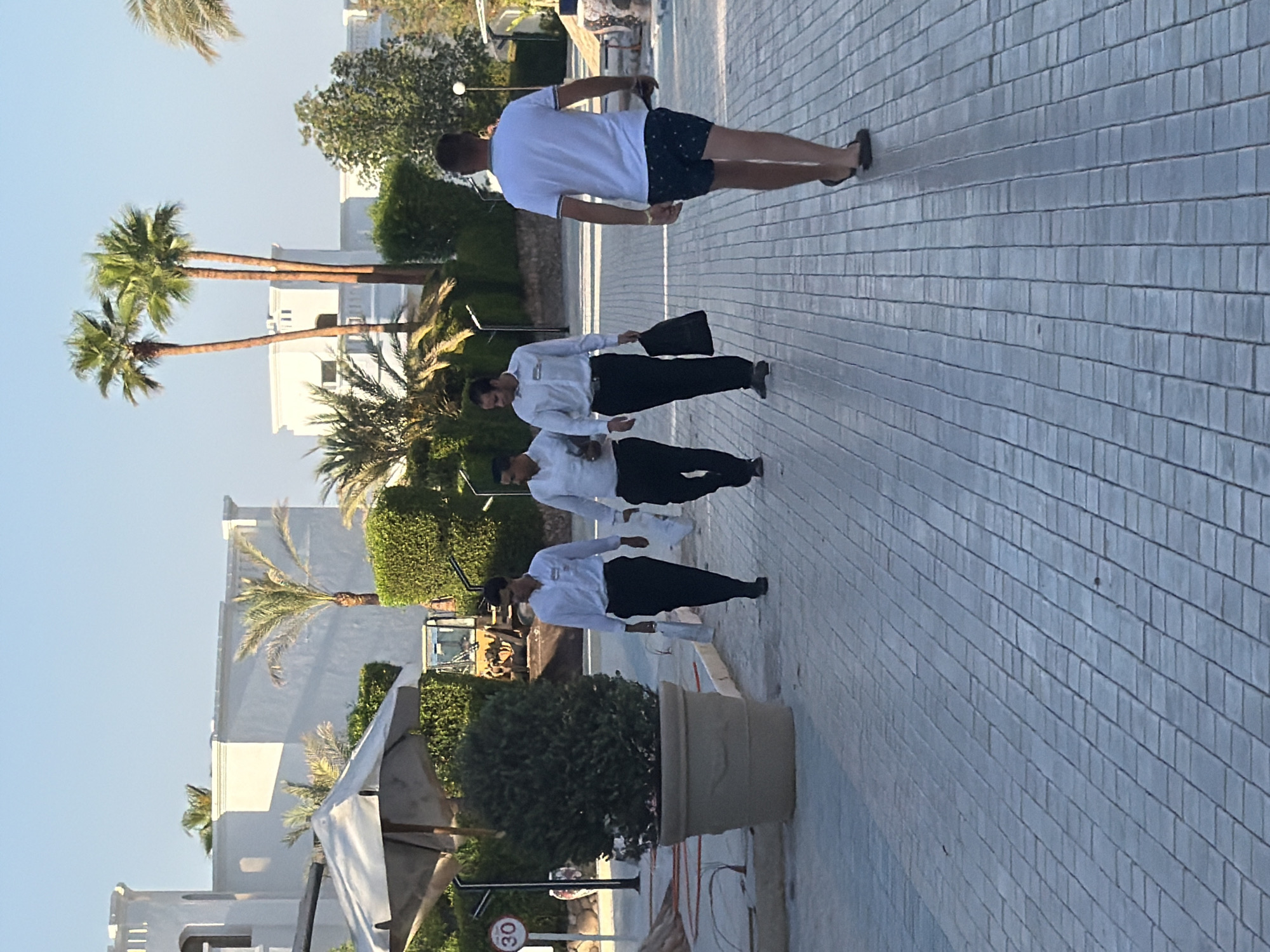 In Sharm El Sheikh, Egypt, on a paved walkway, four individuals are present. The furthest individual from the camera, a male, walks away from the viewer wearing dark patterned shorts and a white t-shirt, with flip-flops. To his left and closer to the camera, three males are walking in the same direction as the first individual. The two males in the foreground of this group are wearing white collared shirts and dark trousers, with one carrying a black bag. The third male in this group, to their right, is also wearing a white collared shirt and dark trousers. Behind these individuals, lush green bushes and palm trees are visible, with white building structures and a yellow construction vehicle in the background. The sky is clear and blue.