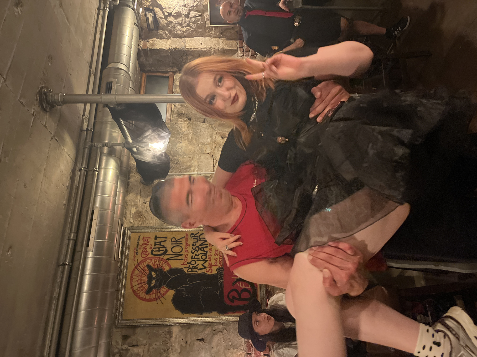 The image depicts an indoor scene with a man holding a woman in his arms. The woman, with red hair, is wearing a black top and skirt. She is smiling and holding up two fingers in a peace sign. The man has a shaved head and is wearing a red shirt. A poster of "Le Chat Noir" can be seen on the wall behind them. Another woman wearing a hat is partially visible in the lower part of the image. In the background, there is a man in black attire and a red scarf. A silver pipe is also visible on the wall.
The location is not identified as Varna, Bulgaria, in the image.
