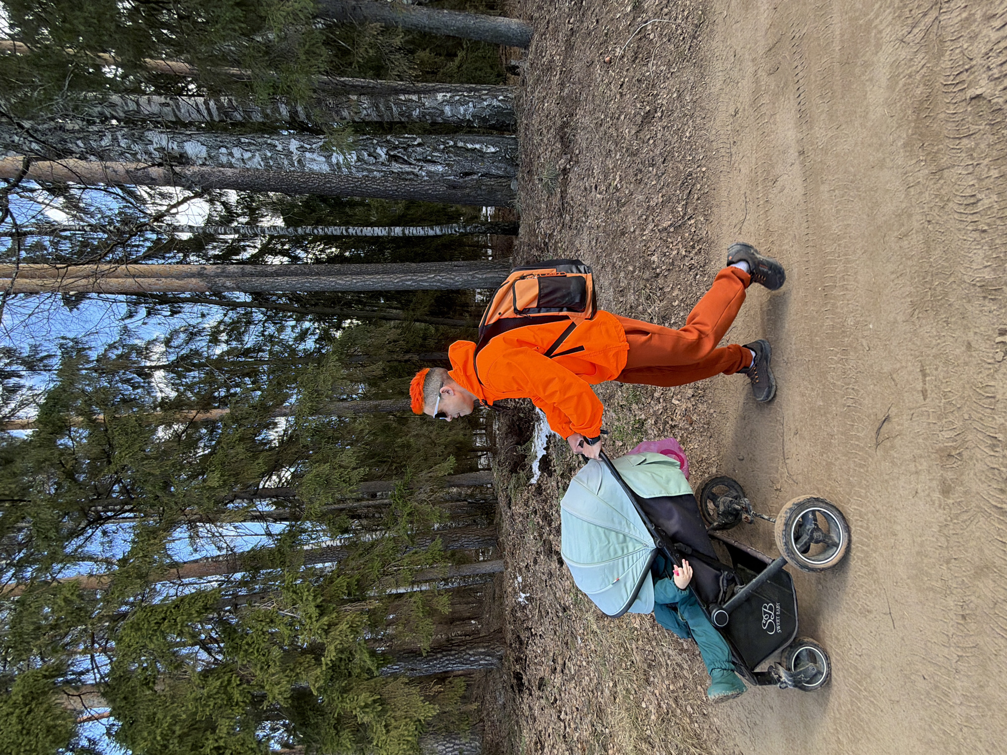 A man in an orange outfit and backpack is pushing a baby stroller on a dirt path in a wooded area. The location is in Chaikovsky, Russia. He is wearing sunglasses and an orange beanie. The stroller is dark and has a light blue hood. The child is visible in the stroller, wearing green and blue clothing. The path is bordered by trees on one side. The sky is visible through the trees, suggesting it's daytime. There are tire tracks visible on the dirt path.