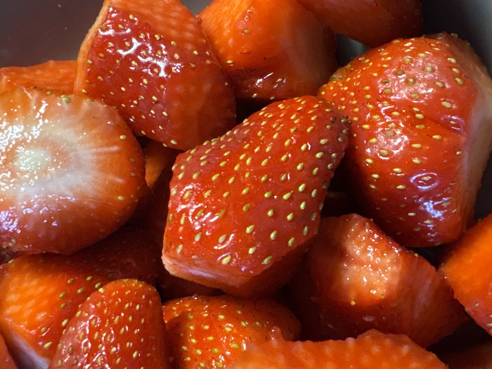 The image presents a close-up, high-angle view of a pile of vibrant red strawberries. The fruit appears to be fresh and glistening with moisture, possibly from being recently washed or sugared. Many of the strawberries are cut, revealing their paler, whitish-pink interiors, while others remain whole or partially intact.
The prominent subjects are the strawberries themselves, filling the frame and creating a rich texture of red and green-yellow achenes (seeds). The achenes are clearly visible across the surface of the fruit, creating a speckled pattern. The scene is tightly cropped, providing an intimate look at the individual berries and their detailed surfaces.
There is no discernible activity taking place beyond the implied preparation or storage of the fruit. The setting appears to be an indoor environment, possibly a kitchen or dining area, given the lack of outdoor elements. The background is dark and out of focus, suggesting the strawberries are contained within a bowl or similar vessel, emphasizing the fruit as the sole focus of the image.
Notable details include the intense, juicy red color of the strawberries, contrasting with the lighter inner flesh and the bright yellowish-green of the achenes. The reflective sheen on the fruit indicates freshness and moisture. There are no visible people, specific location cues (other than the implied indoor setting), or text within the image. The time of day or weather conditions are not apparent from the photograph. While the location context is provided as Saint Petersburg, Russia, there are no visual elements in the image to confirm this.