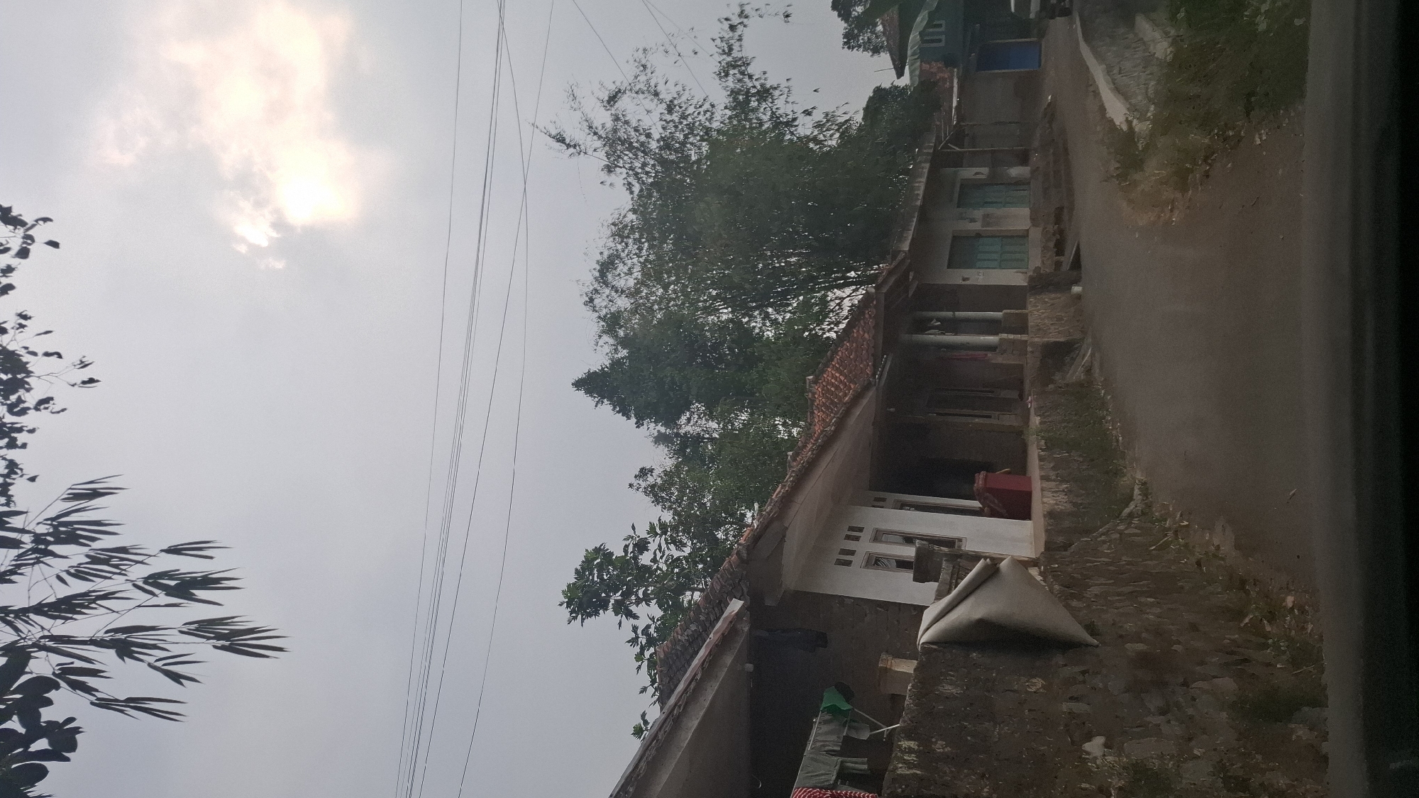 A low-angle, wide shot captures a quiet street in the village of Sukasari, Indonesia, under an overcast sky. The road, paved with asphalt, curves gently to the right, bordered by a low, stone retaining wall on the left and a grassy embankment on the right. Along the left side of the road, a series of attached, single-story homes with terracotta tile roofs are visible. The architecture is simple, with some homes featuring white facades and dark-framed windows. Laundry hangs on a clothesline in front of one house. Tall bamboo stalks and other lush green trees rise behind the buildings, suggesting a rural or semi-rural setting. The sky is a muted grey with a patch of diffused sunlight breaking through the clouds on the upper right. Power lines crisscross the upper portion of the frame, silhouetted against the sky. The foreground is obscured by the dark interior of what appears to be a vehicle. No people are present in the scene.