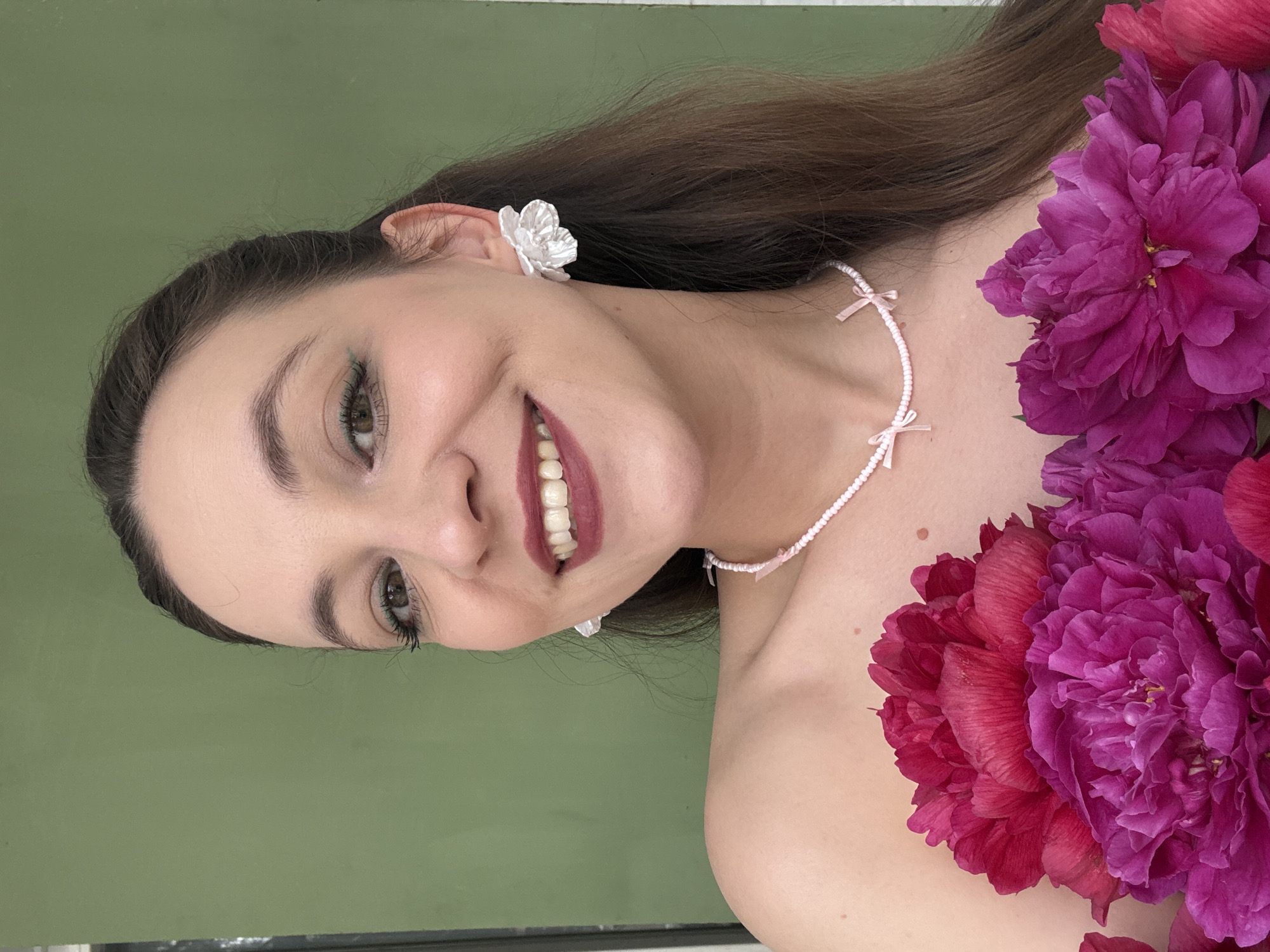This is a portrait of a young woman with long, dark brown hair and brown eyes, who is smiling. She is wearing a white flower-shaped earring and a delicate necklace with small pink bows. The woman is adorned with large, vibrant pink and red peonies, which frame her face and chest. The background is a solid, muted green color. The image appears to be taken indoors, possibly in a studio setting, given the controlled lighting and uniform background. The woman's expression conveys happiness and a sense of playful elegance. The overall aesthetic suggests a whimsical or romantic theme. There is no visible text in the image.