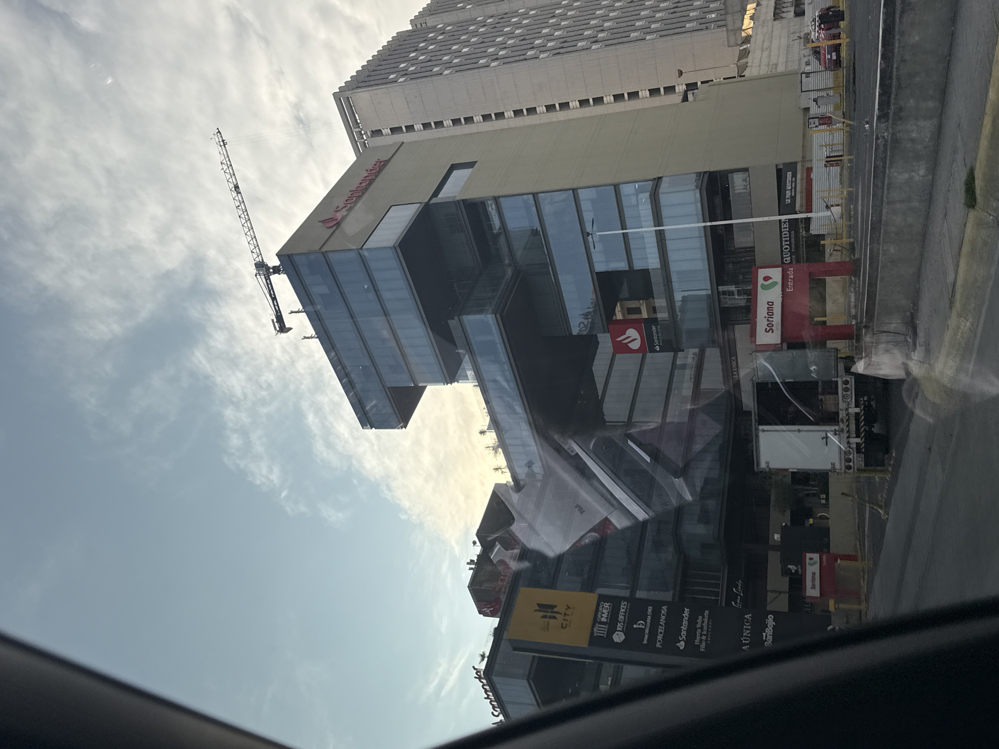 The photograph captures a view of a commercial complex in Heroica Puebla de Zaragoza, Mexico. The architecture is modern, incorporating glass and steel in its design. Prominent signage indicates that the complex houses a Santander bank branch and a Soriana supermarket. Other businesses and services are also visible on signs. A construction crane is present near the buildings, suggesting ongoing or recent construction activities. The photograph is taken from inside a vehicle, with the car window visible in the foreground.