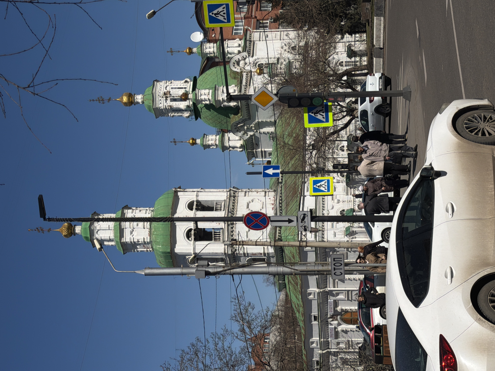 This upright image captures a vibrant street scene in Krasnoyarsk, Russia, under a clear blue sky. The prominent feature is a magnificent white building with intricate architectural details, topped with multiple green domes and golden spires, characteristic of an Orthodox church or cathedral. Bare tree branches, indicative of late winter or early spring, frame the upper left corner and line the street.
On the right side of the frame, a street with asphalt pavement is visible, hosting several vehicles. A white car, possibly a sedan, is prominently parked in the foreground, partially obscuring a group of pedestrians on the sidewalk. Further down the street, another white SUV or crossover and a red car are visible.
Several people, dressed in jackets and coats, are gathered on the sidewalk, some appearing to wait near a pedestrian crossing. Overhead wires crisscross the sky. The street is equipped with various signs and infrastructure: a traffic light showing a green signal, multiple pedestrian crossing signs (blue square with a white pedestrian icon), a yellow diamond-shaped sign indicating a priority road, a blue rectangular sign with a white arrow pointing left (one-way street), and a red circular "no stopping" sign with a blue background and a red cross. Another sign depicts a camera and a tow truck icon, likely warning of enforcement or surveillance.
A square white sign with black text reads "СТОП" (STOP). On the facade of a building in the background, text "ТРИБУНА" and what appears to be a phone number "1988881" are visible. The bright lighting and sharp shadows suggest a sunny day, likely mid-day.