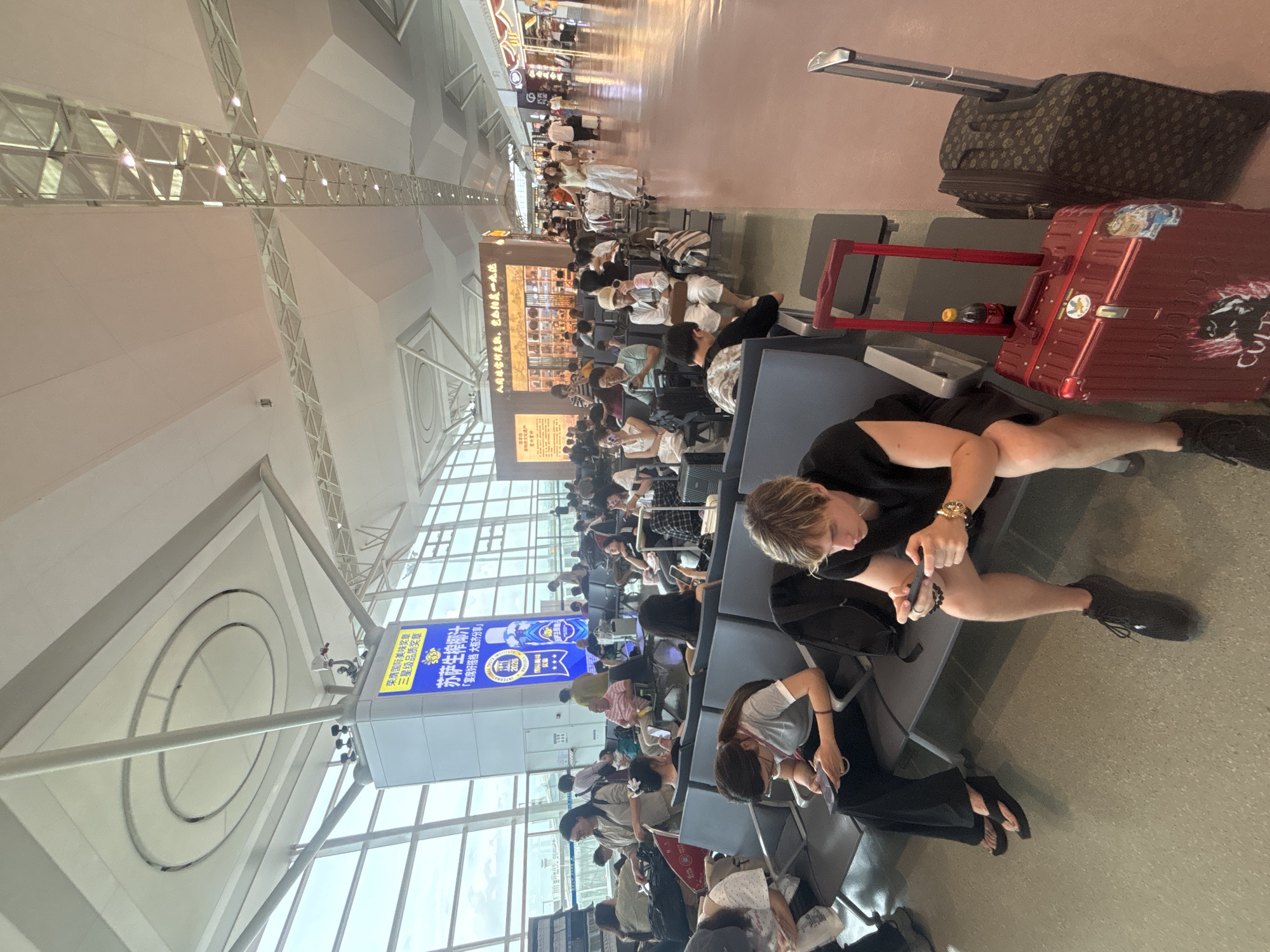 The image depicts an indoor waiting area, likely an airport terminal, in Xinwu District, China. Numerous individuals are seated on gray benches, appearing to wait for flights or transportation. Many are engaged with their mobile phones. In the foreground, a young man with blond hair is seated, engrossed in playing a game on his phone, with a red rolling suitcase beside him. To his right, another dark-colored rolling suitcase stands. The environment features large windows allowing natural light, a high ceiling with exposed structural elements and lighting, and a polished floor. Several signs with Chinese text are visible on a pillar and a wall in the background, indicating the location is likely within China. The overall atmosphere is one of quiet waiting and individual engagement.