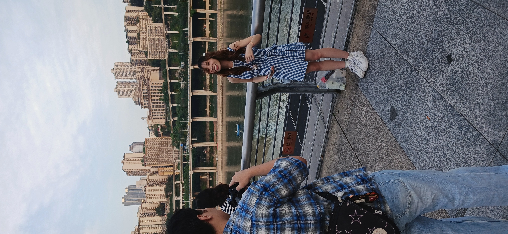 The image depicts a scene in 江北城街道, China, where a person is photographing a young woman posing by a railing. The woman is wearing a blue and white checkered dress and white sneakers. She is smiling and has her arms slightly raised. Behind her is a body of water and a cityscape with multiple buildings and bridge structures. The photographer is wearing a blue plaid shirt and light-wash jeans, holding a camera. He is partially visible in the foreground, with another person behind him. A plastic bag with some items is placed on the ground near the woman. The scene appears to be outdoors, on a paved walkway.