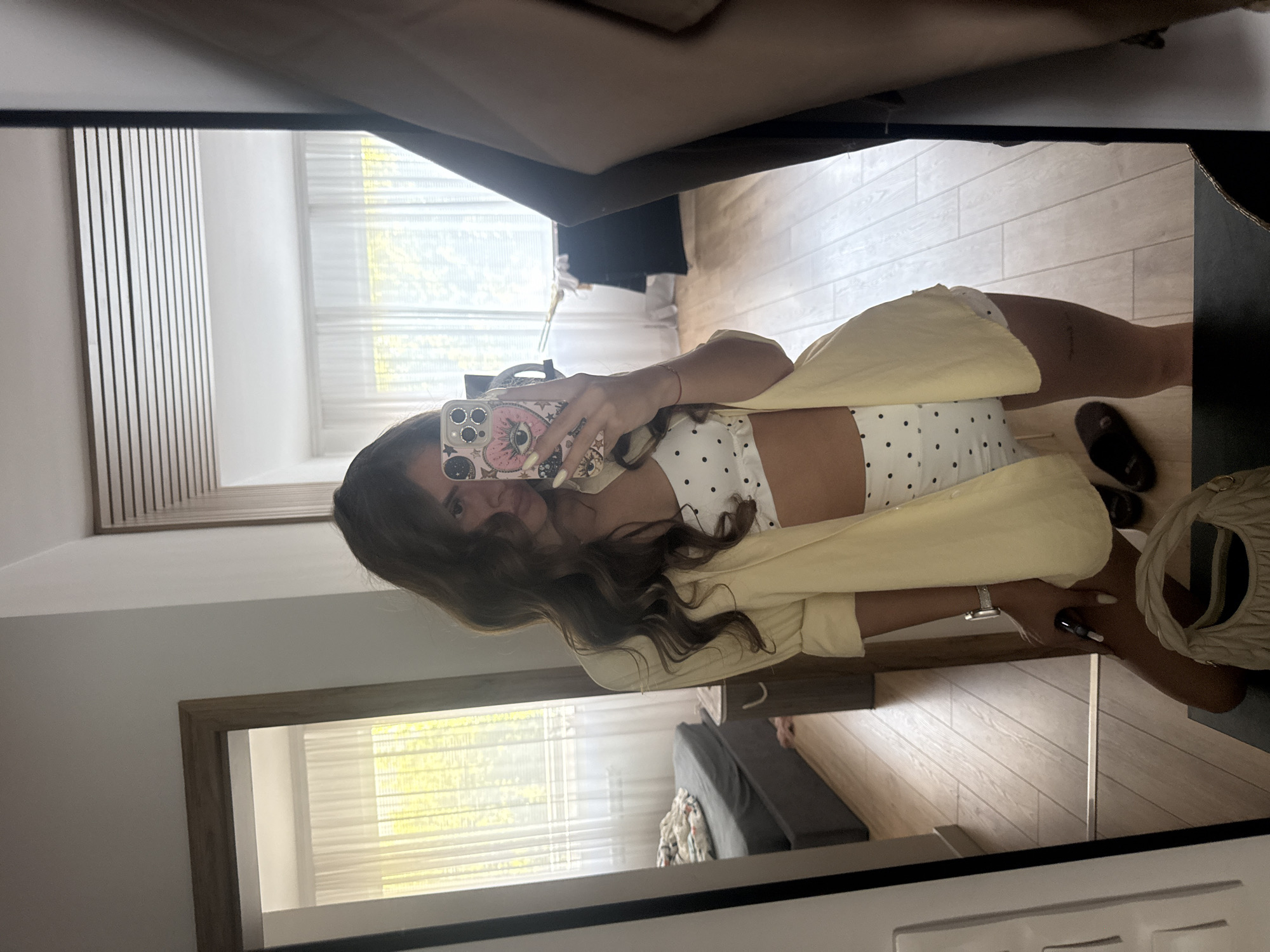 The image shows a young woman taking a mirror selfie in what appears to be a bedroom. She is wearing a white cropped top with black polka dots, white shorts, and a pale yellow open cardigan. She has long, dark, wavy hair that partially covers her face. Her left hand is holding a smartphone, which has a pink case with a large eye and other cartoonish designs. She is wearing a silver watch on her left wrist and has painted fingernails. Her right arm is resting on her hip.
The setting is a room with light-colored wooden floors. In the reflection, we can see a large mirror, part of a bed with bedding, and what appears to be a window with white blinds covered by sheer curtains. There is also a glimpse of a wooden door frame. To the right, a portion of a cream-colored, ruched handbag is visible. The lighting suggests it could be daytime, possibly late morning or afternoon, given the brightness coming from the window. The overall mood is casual and relaxed. The image is taken indoors, within a residential setting.