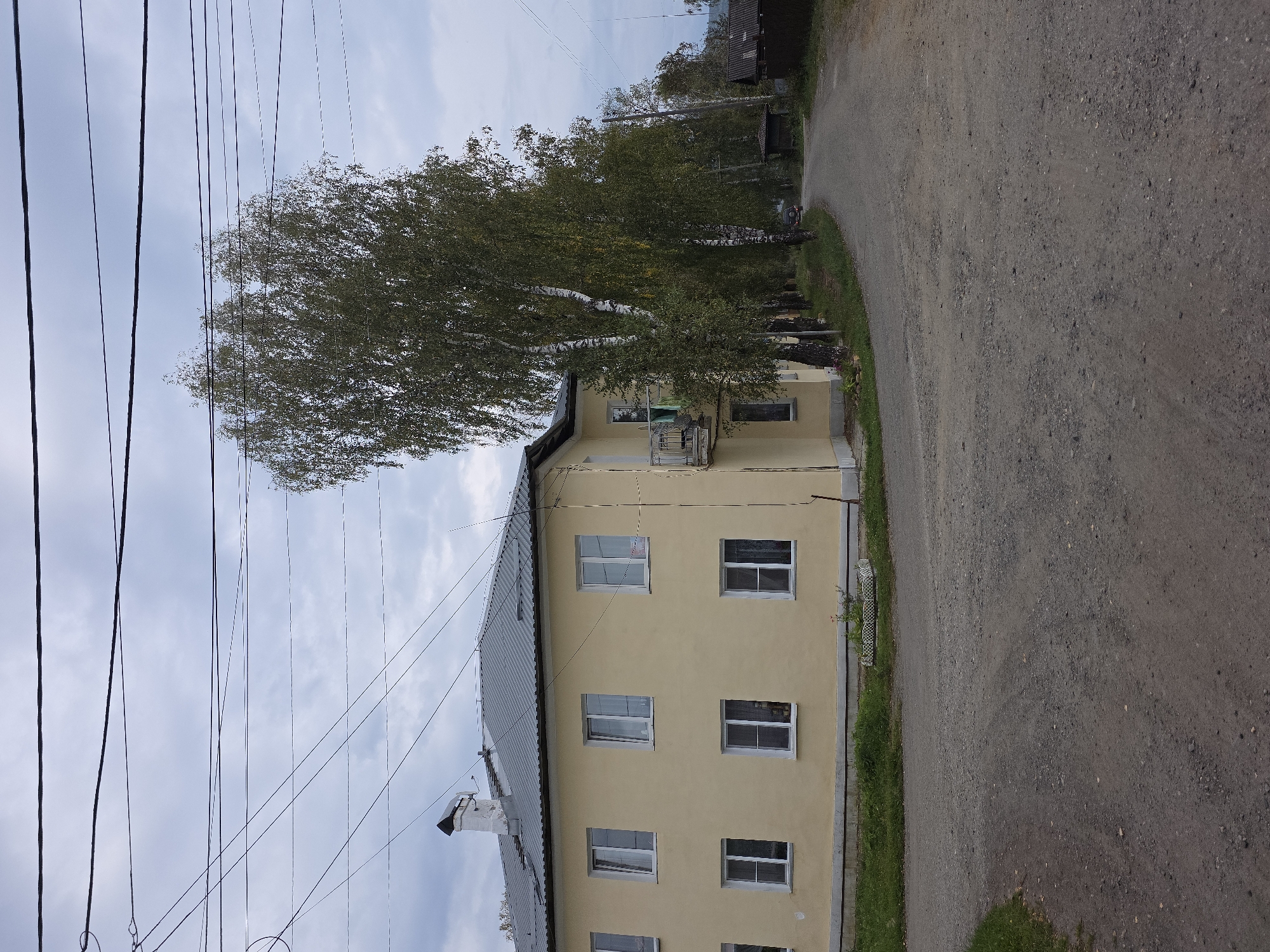 This is a medium shot, eye-level, outdoor photograph of a two-story, pale yellow apartment building situated on a gravel road in Magnitka, Russia. The building has a grey, corrugated metal roof and features six white-framed windows on each floor. A small balcony with a white railing is visible on the second story. Several large birch trees with white bark line the side of the building and the road. A white chimney with a dark cap rises from the roof. The road is unpaved and appears to be in a rural or semi-rural setting. The sky is overcast with a light grey color, suggesting a cloudy day. In the distance, a dark-colored vehicle is parked on the side of the road. Two small, dark-colored outbuildings or sheds are also visible further down the road. The scene is devoid of any people.