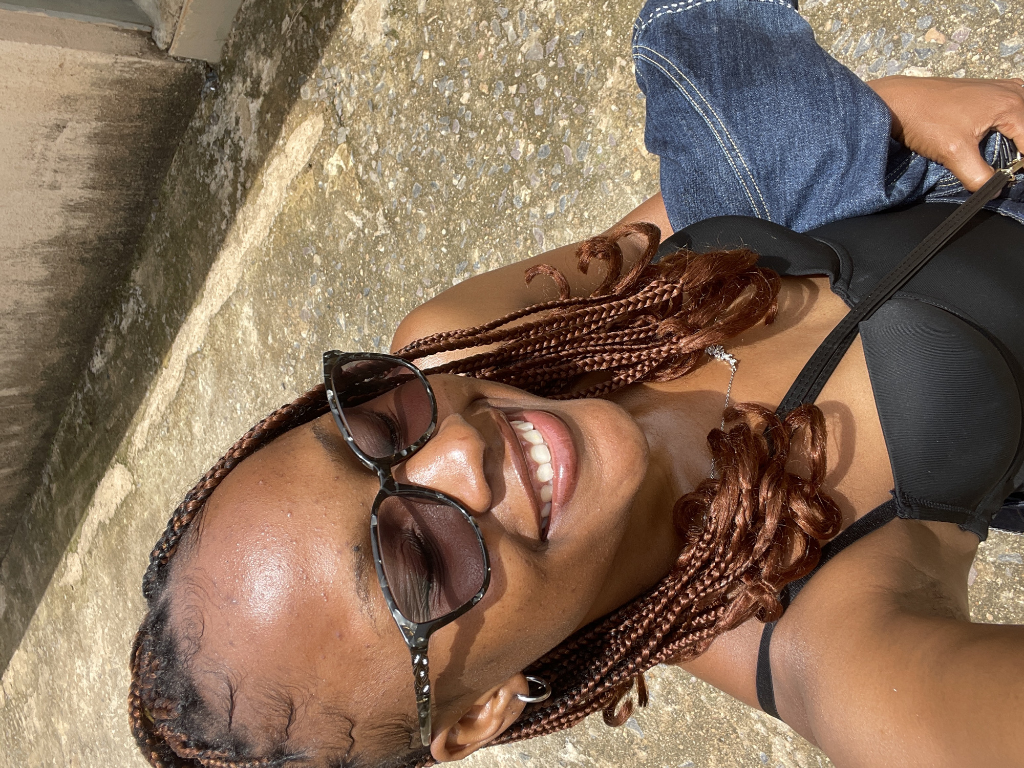 A woman with braided hair and sunglasses smiles while looking down. She is wearing a black top and holding blue denim clothing. The background appears to be an outdoor area with a concrete or stone ground and a wall. The lighting suggests it is daytime. The location in the city of Rafin Par, Nigeria is indicated by the context provided.