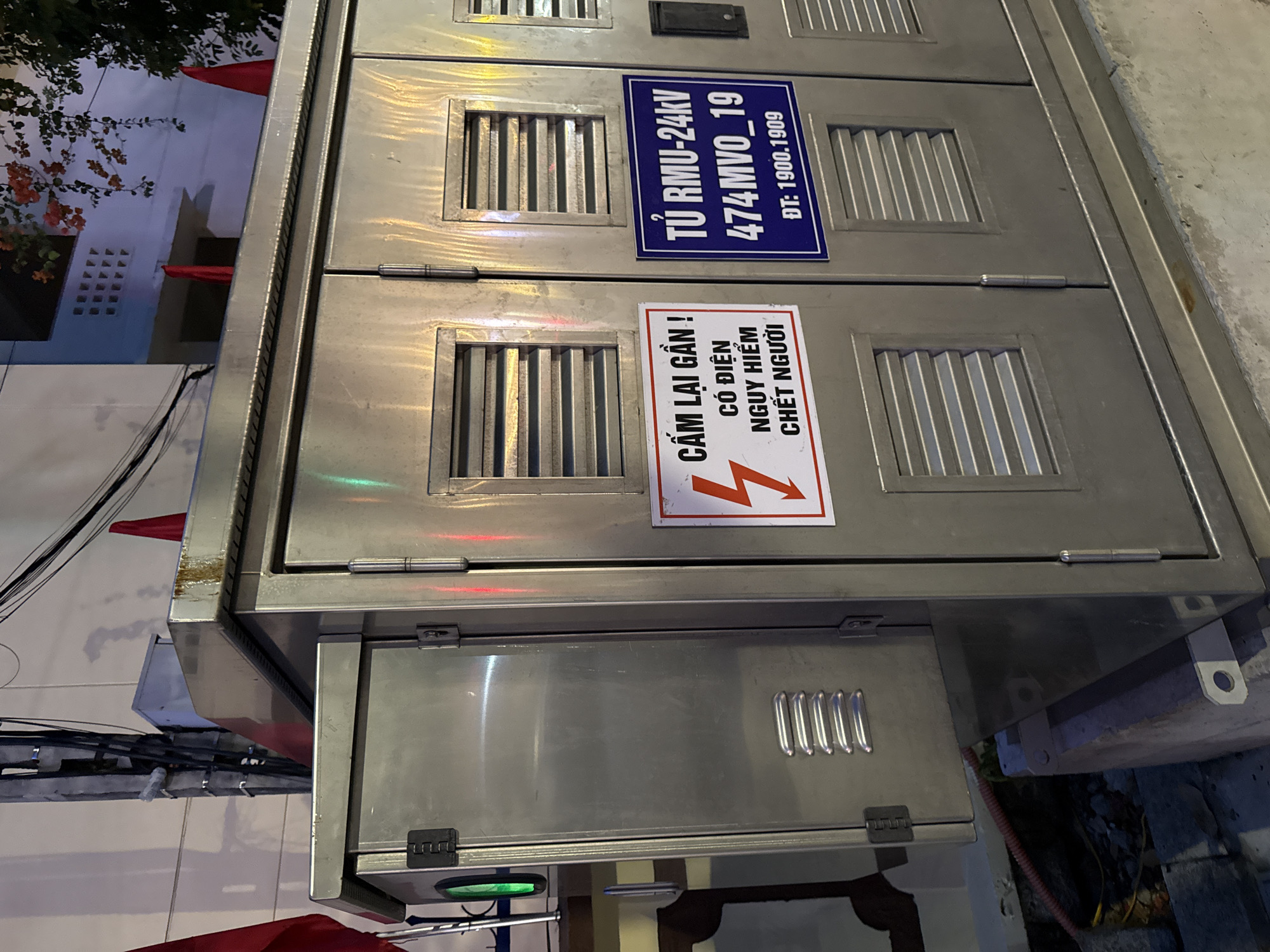 The image shows a metal electrical cabinet located outdoors in Nha Trang, Vietnam. The cabinet has multiple doors with ventilation grates. Two warning signs are visible on the front of the cabinet. One sign, with a red lightning bolt symbol, reads "CẨM LẠI GẦN! CÓ ĐIỆN NGUY HIỂM CHẾT NGƯỜI," which translates to "DO NOT APPROACH! DANGER HIGH VOLTAGE DEATH." The second sign, on a blue background, reads "TỦ RMU-24KV 474MVO_19 ĐT: 1900.1909," indicating it is a 24kV RMU cabinet with a specific identification number and a phone number. Red flags are visible in the background, suggesting a possible holiday or event. The environment appears to be a street or public area, with electrical poles and wires also present. The lighting suggests it is nighttime or dusk.