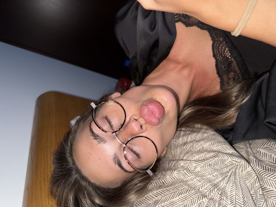 A woman lies sideways in bed, eyes closed and lips puckered, wearing round tortoiseshell glasses. She rests on a patterned pillow with a black silky top trimmed with lace around the neckline. The top of her shoulder and upper arm are visible in the upper right corner. A tan elastic hair tie is on her arm. The background features a dark ceiling, a pale wall, and a corner of wooden furniture.