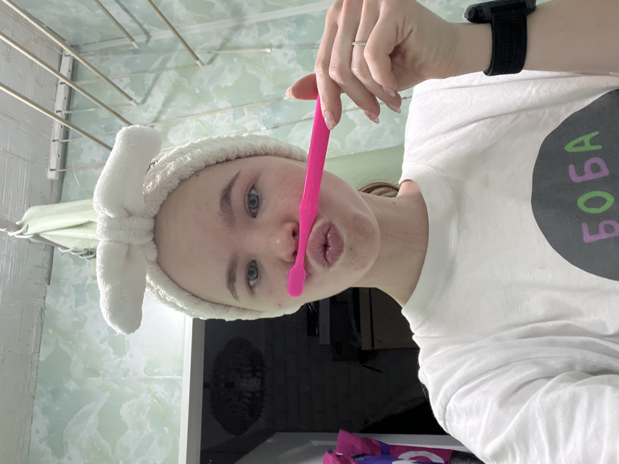 A woman with fair skin, light brown hair and blue eyes wears a white, plush headband with a bow. She holds a pink utensil vertically to her lips, puckering her mouth. She wears a white t-shirt with a black circle containing the word "BOBA" in alternating green and purple letters. She wears a black wrist watch on her left wrist. The background shows a green and white tiled wall and a metal drying rack. A partial view of a bathroom counter is visible in the lower center. The location is identified as Chaikovsky, Russia.