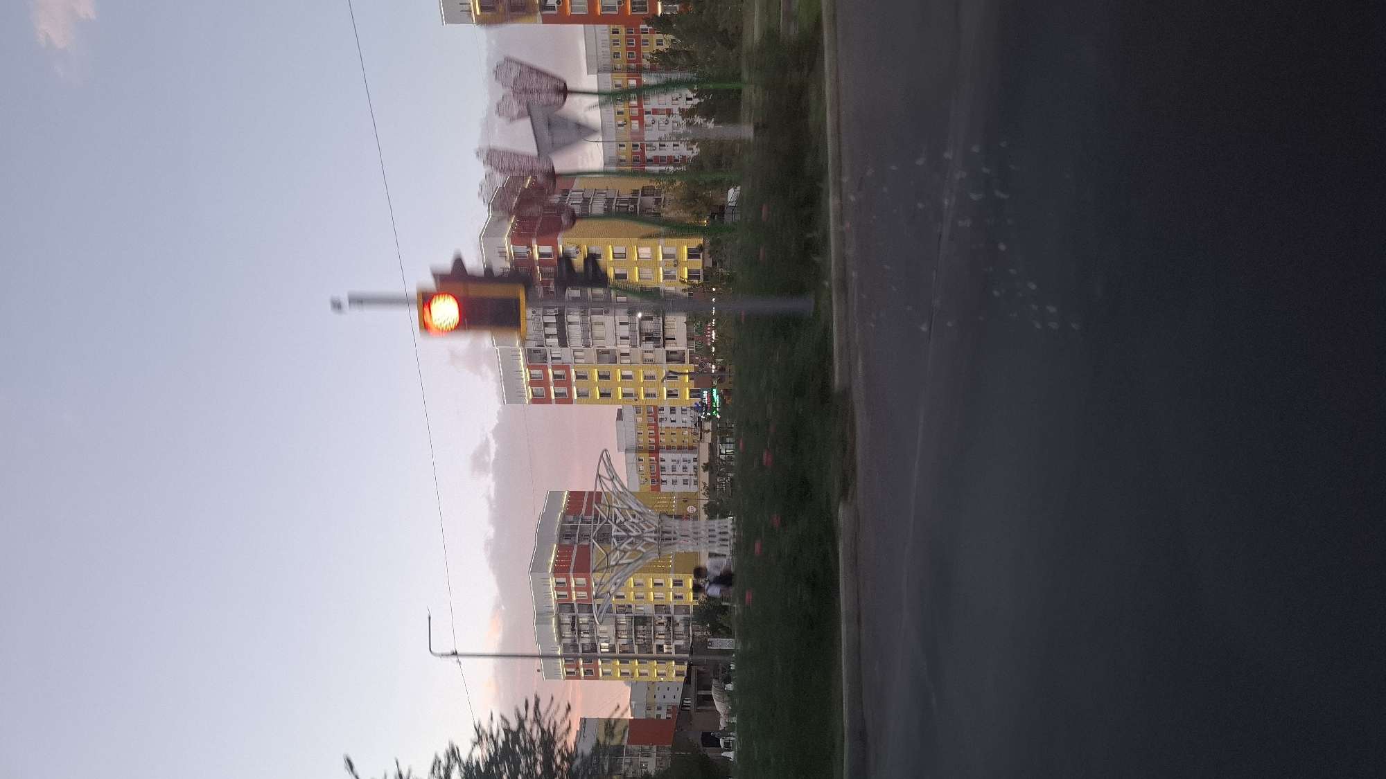 The image depicts a street view in the city of Shymkent, Kazakhstan, likely during dusk or twilight. The sky is a soft gradient of pink and pale blue, with some scattered clouds. In the background, several brightly colored apartment buildings stand tall, their windows illuminated. The buildings are primarily yellow and red with white trim. In the foreground, a street with dark asphalt is visible. A traffic light with a red signal is prominent on the right side of the frame, appearing slightly blurred due to motion. To the left of the traffic light, there are decorative sculptures resembling large flowers. In the middle ground, a white, tree-like sculpture with an intricate design stands in a landscaped area. A few people are walking in the distance, near the base of the tree sculpture. Lush green bushes and some flowering plants form a border between the street and the landscaped area. A street lamp can be seen on the far left. The overall impression is of a modern urban environment with artistic embellishments, captured during a transitional time of day.