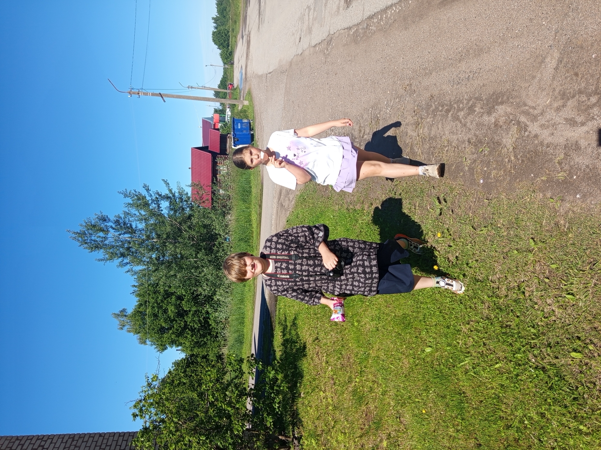 Two children are walking on a dirt road in a rural area. The child on the left is wearing a dark patterned shirt, dark shorts, and sneakers. They have a camera around their neck and are holding a package of snacks. The child on the right is wearing a white t-shirt with graphics, a purple skirt, and sneakers. They are eating an ice pop. In the background, there are trees, houses with red roofs, and a blue dumpster. The sky is clear and blue. The location is Gdov, Russia.