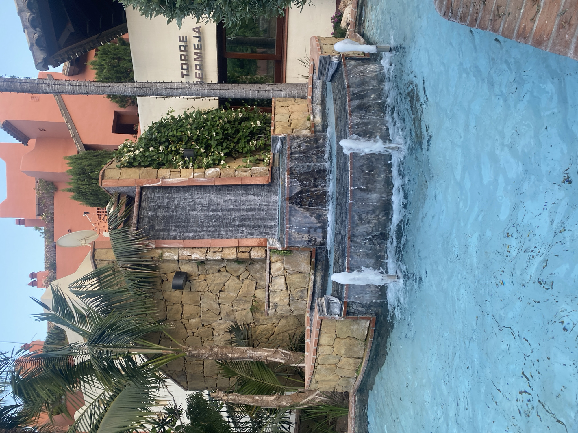 The image depicts a multi-tiered water feature with a flowing fountain, set against a backdrop of a building and lush greenery. The water feature is constructed with stone and brick, with water cascading down several levels. Jets of water are actively shooting from pipes, creating a dynamic visual effect.
In the background, a building with an orange facade and tiled roof is visible, along with a cream-colored section bearing text that reads "TORRE BERMEJA". The building appears to be modern with traditional architectural elements. Several palm trees and other green plants, including a vine with white flowers, are interspersed around the water feature and building, suggesting a warm climate and possibly a resort or residential setting. The sky is visible at the top of the image, appearing clear and light, indicative of daytime.
The scene suggests a serene and relaxing environment, possibly a public space or a private garden area. The activity is the natural movement and sound of the water, enhanced by the fountain's jets. There are no people visible in the image. The overall impression is one of a well-maintained outdoor space designed for aesthetic appeal and enjoyment. The location context of Estepona, Spain, aligns with the Mediterranean architecture and vegetation.