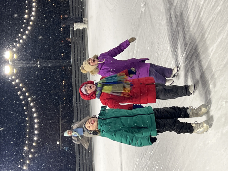 An outdoor ice rink in Saint Petersburg, Russia, is depicted at night during a snowfall. The scene is illuminated by overhead floodlights and strings of decorative globe lights. Three children, aged approximately 6-10 years, are centrally positioned on the ice, all wearing winter coats, pants, beanies, and ice skates. From left to right:
1. A child in a green puffer jacket, black pants, and a striped beanie extends their tongue upwards, looking towards the falling snow.
2. A child in a red puffer jacket, dark pants, a multi-colored striped scarf, and a white beanie has their mouth open wide, looking upwards, as if shouting or catching snow.
3. A child in a purple puffer coat, purple pants, and a beige pom-pom beanie also has their mouth open wide, looking upwards, with their right arm slightly extended.
The ice surface shows skate marks. In the background, an adult in a light blue top and brown coat stands further back on the ice with arms raised. To the far right, another person in white clothing is partially visible near a dark barrier or fence. Snowflakes are visibly falling and are illuminated against the dark sky.