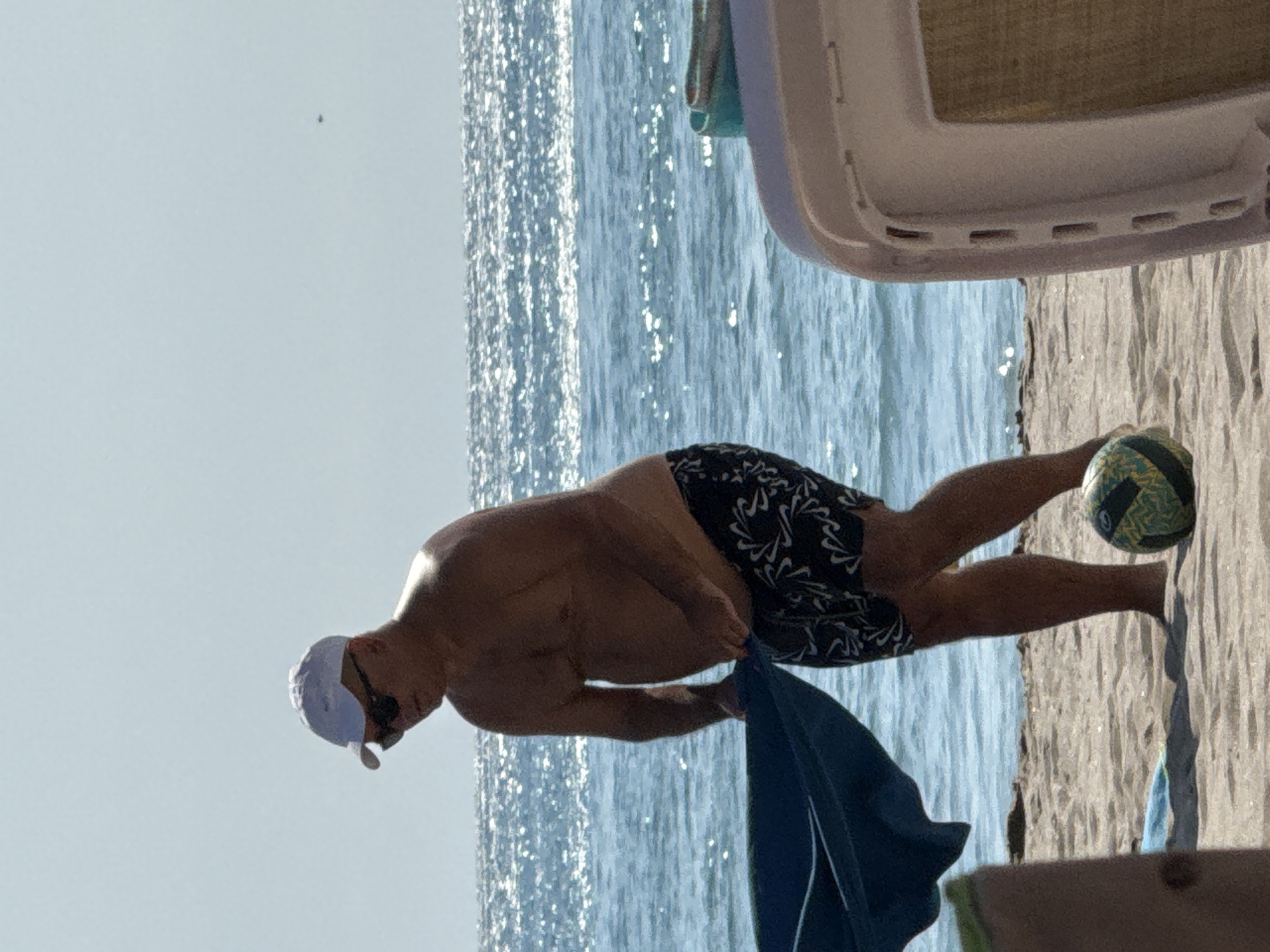A man, wearing a white baseball cap, sunglasses, and black and white patterned swim trunks, stands on a sandy beach. He is holding a large, dark blue towel or blanket. Next to his feet is a multicolored ball. In the background, the blue ocean shimmers under a clear sky, with white caps on the waves. To the right of the man, a portion of a white beach chair is visible. The scene is outdoors on a sunny day.
