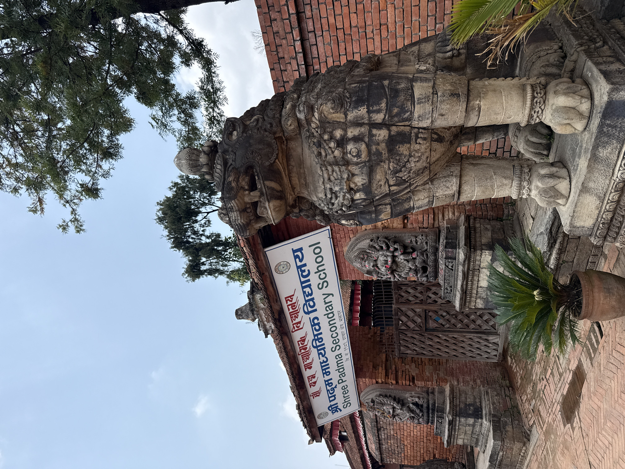 The image shows the entrance to Shree Padma Secondary School in Bhaktapur, Nepal. A large stone lion statue stands guard. The school sign, visible in the lower center, features the school name in both Nepali and English. Behind the lion and sign is a brick building with ornate wooden doors and carvings. Potted plants are visible. Tree branches are visible above and to the left. The sky is pale blue with some clouds.