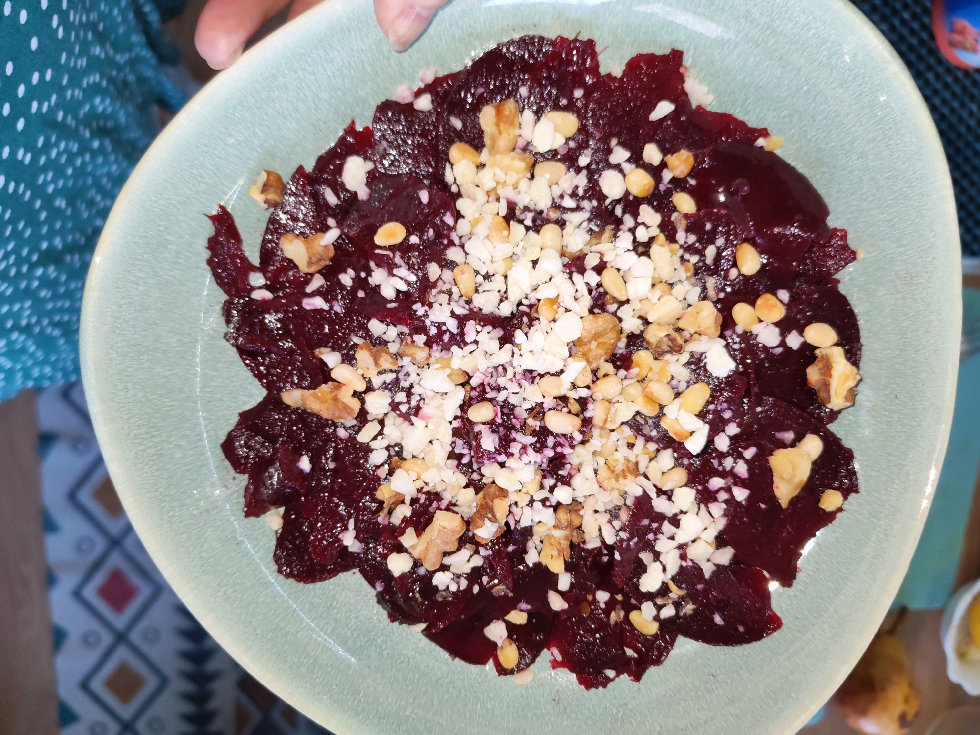 A close-up, top-down shot captures a person holding a light blue-green, irregularly shaped ceramic plate filled with a vibrant beet dish. The main subject is the food, consisting of thinly sliced, dark red-purple beets spread across the plate, resembling a carpaccio or salad. This base is generously topped with a mix of chopped walnuts, lighter-colored pine nuts, and a significant amount of a white crumbled ingredient, possibly feta cheese or a similar soft cheese, creating a visually appealing contrast of colors and textures.
Only the fingers and thumb of the person holding the plate are visible at the top center of the frame. The person is wearing clothing made of a teal or dark green fabric patterned with small white polka dots, visible on the left side. The background to the bottom left features a blurry patterned mat or tablecloth with geometric diamond shapes in shades of blue, red, brown, and white. Further in the blurry background on the top right, hints of other objects, possibly a yellow container, are discernible.
The scene suggests an indoor, domestic setting, likely a kitchen or dining area, where a dish is being presented or prepared. The lighting is bright and even, accentuating the details of the food. The image is devoid of any visible text. The context for this scene is Izhevsk, Russia.