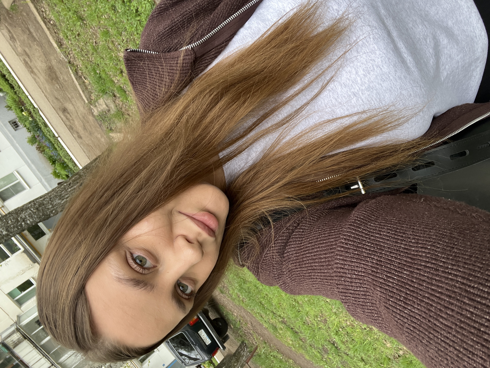 A woman with long brown hair and green eyes is facing the camera, positioned in a natural outdoor setting. She is wearing a white t-shirt and a brown zip-up jacket. In the background, a green lawn and a building with windows can be seen, suggesting an urban residential area. A black car is visible in the lower part of the frame. The license plate number of the car is O745TX52. Based on the Cyrillic license plate and the style of the buildings, the location may indeed be Chaikovsky, Russia, or another location in Russia.