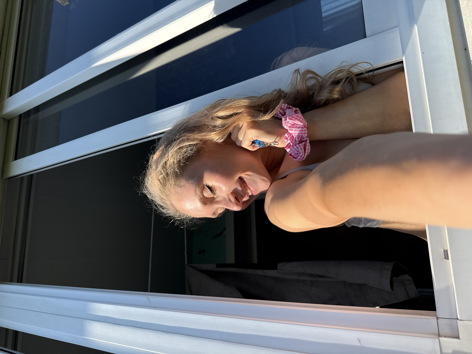 The image shows a young woman with blonde hair, wearing a grey tank top and a pink and white checkered scrunchie on her wrist, leaning out of an open window. She is smiling with her eyes closed and her head tilted. The window frame is white. In the background, dark curtains are visible, along with a dark interior space. The lighting suggests it is daytime, possibly late afternoon, with sunlight illuminating the woman and the window frame. There is no discernible information within the image to indicate the location in the city of Perm, Russia.