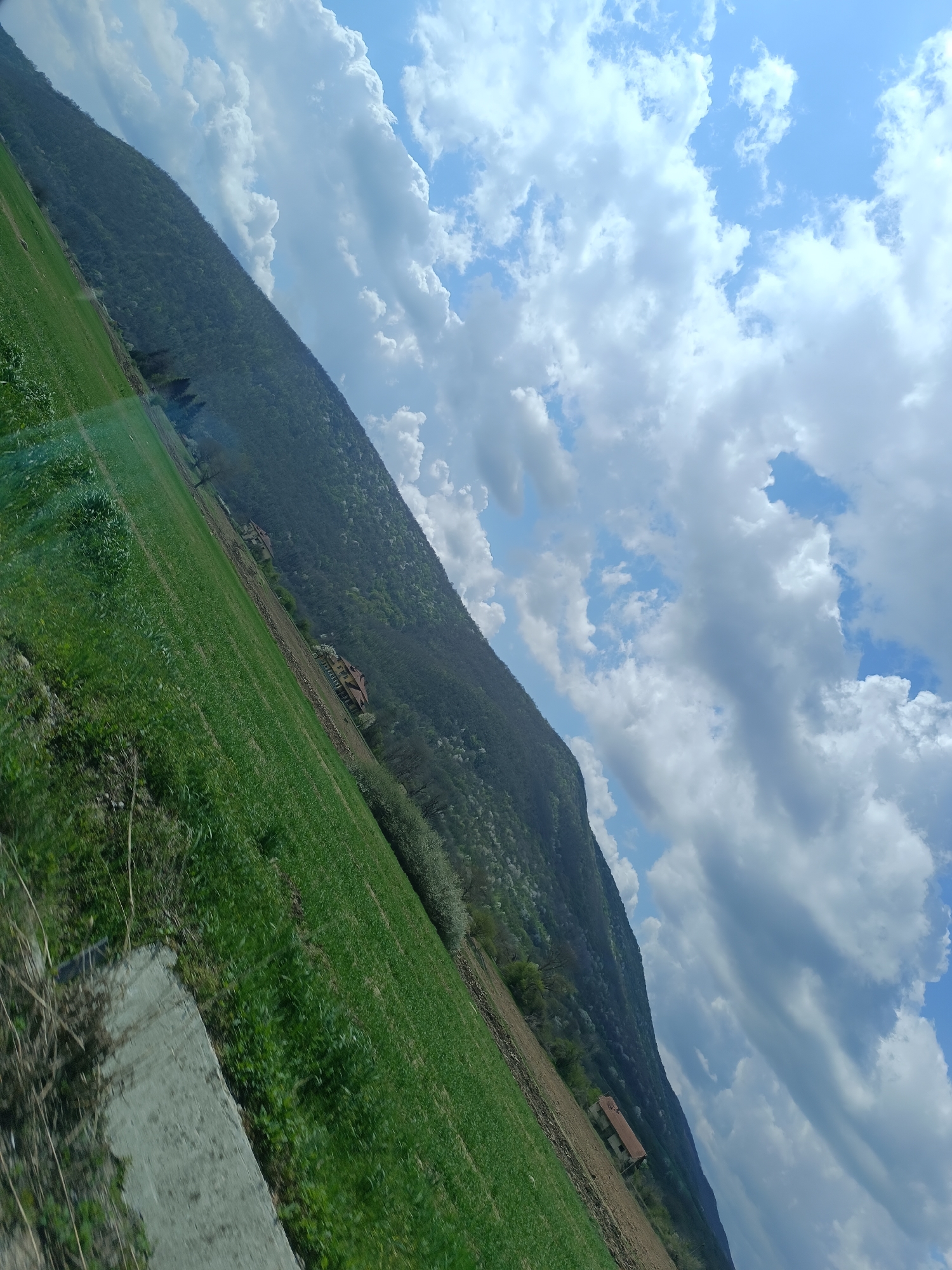 The image shows a landscape of a green hillside with a mountain in the background under a sky with scattered clouds. There is a concrete structure in the foreground, possibly a curb or a small wall. Scattered trees and shrubs grow on the hillside. Several buildings are situated on the hillside, further up toward the mountain. The photograph is taken at an angle. The location is Unknown, Unknown.