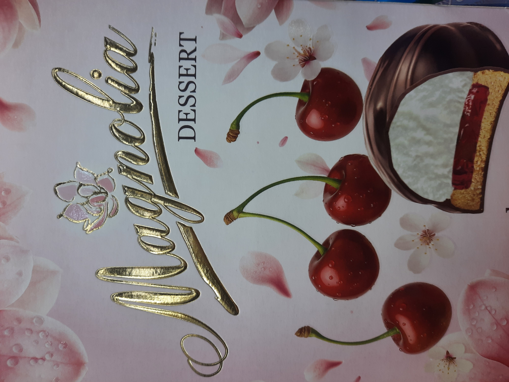 The image is a close-up shot of the packaging for a dessert, likely a cake or pastry. The dominant text on the packaging reads "Magnolia DESSERT" in a stylized golden font. The background features a soft pink hue with illustrations of magnolia flowers and petals, as well as several glossy red cherries with stems. In the lower right corner, a cross-section of the dessert is visible, revealing layers of chocolate coating, white filling, and a red center, all on top of a biscuit base. The packaging appears to be made of a material with a slight sheen, reflecting light in places. The image does not depict any specific location in the city of Парэчча, Belarus, as it is a product shot.