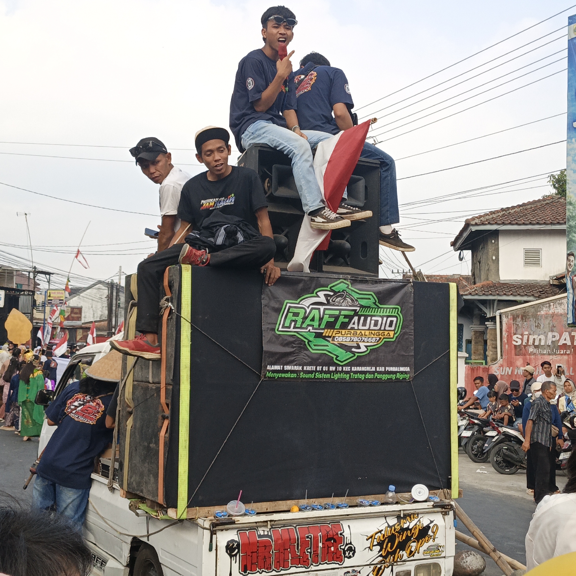 The image shows a group of young men on top of a truck with a large speaker system in what appears to be a street scene in Randudongkal, Indonesia. One man in the center is holding a microphone and singing, wearing sunglasses and a dark blue t-shirt. Another man to his left, wearing a black t-shirt and a hat, is looking towards the microphone. To the right of the singer, a man in a dark blue t-shirt is sitting with a red and white Indonesian flag.
The truck has a large black speaker box mounted on its back, with a banner that says "RAFF AUDIO PURBALINGGA". The truck itself is white and has graffiti-style lettering on the rear, including the words "MR. MELLTLE" and "Tulusmu wingi opo?".
In the background, there are buildings, power lines, and a crowd of people, many of whom are on motorcycles. Some of the people in the crowd are holding up phones, likely recording the event. The overall atmosphere suggests a public gathering or celebration. The clothing of the people and the style of the buildings are consistent with a Southeast Asian setting.