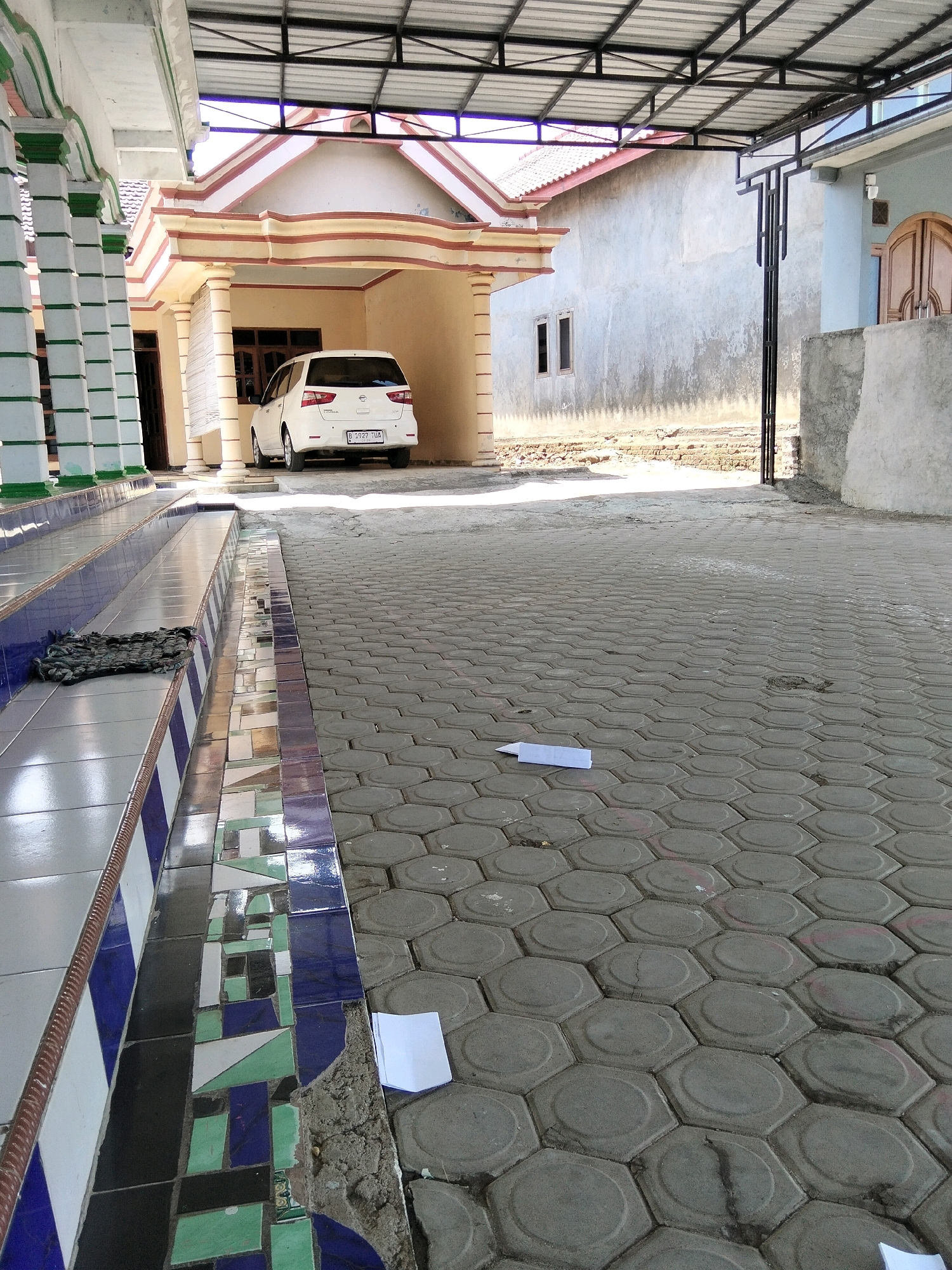 The image depicts an outdoor scene in Jenangan, Indonesia, likely a residential area or a commercial establishment. In the foreground, a patterned tiled walkway transitions to hexagonal concrete paving stones. A white van is parked under a covered carport in front of a building with cream-colored walls and a red-tiled roof. To the left, a covered walkway with white columns, accented with green and blue, leads to an entrance. A dark-colored doormat rests on the tiled steps of the walkway. The background shows a grey wall of a building and a glimpse of a wooden door with arched panels. Scattered on the paving stones are two folded pieces of white paper. The lighting suggests it is daytime, with sunlight casting shadows.