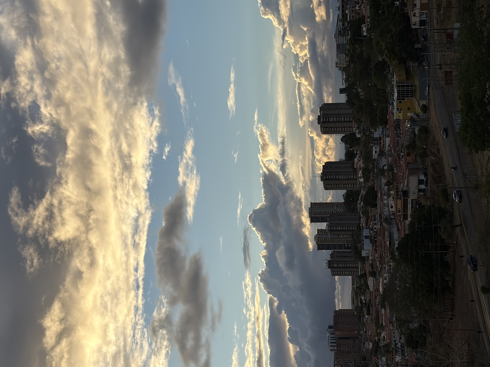 An elevated panoramic view depicts a cityscape in Ciudad Guayana, Venezuela, at an estimated late afternoon or early evening, characterized by dramatic cloud formations and indirect sunlight. The upper two-thirds of the image are dominated by a sky featuring extensive cumulonimbus clouds, brightly illuminated with golden hues from above, contrasting with pockets of clear blue sky and darker cloud masses closer to the horizon.
The lower third of the image comprises the urban environment. In the mid-distance, a cluster of approximately seven to eight multi-story buildings, appearing as dark high-rises or apartment blocks, are visible, partially silhouetted against the bright clouds. Below and extending into the foreground is a dense residential area composed primarily of numerous low-rise buildings with predominantly orange or red-tiled roofs, interspersed with mature trees.
A multi-lane asphalt road, likely an avenue, curves through the lower mid-ground from left to right. Several vehicles are observed on this road: a dark sedan in the left lane and another dark sedan further along the right lane. A white sedan is parked off the road near a distinctive yellow building with a grid-patterned facade, located in the mid-right. A motorcycle is also visible on the road's left side. Scattered individuals are visible walking alongside the road: two on the left verge and one near the dark sedan on the right. In the far left foreground, a tall metal lattice structure, consistent with a power transmission tower, is prominent. Electrical poles and wiring are also visible on the right side of the road.