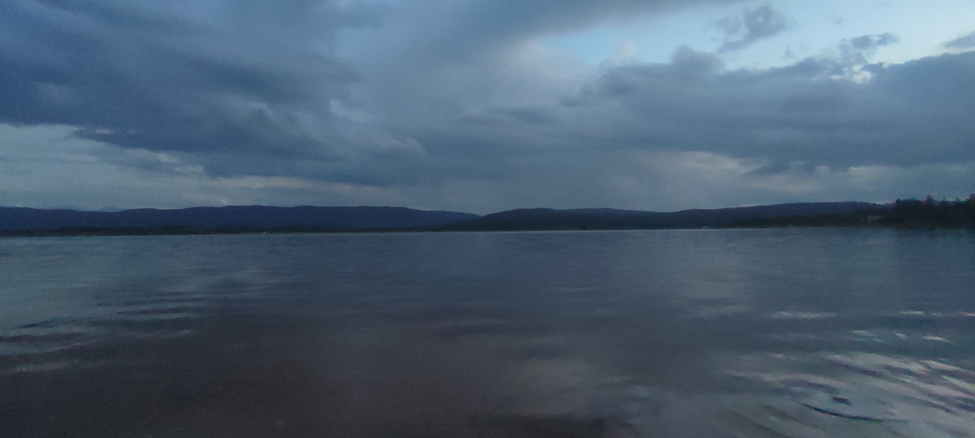 The image presents a tranquil, wide landscape featuring a large body of water under a dramatic, cloudy sky. The foreground and midground are filled with the calm surface of a lake or reservoir, rendered in shades of dark blue-gray, reflecting the overcast sky and showing subtle ripples.
In the middle distance, a long, dark silhouette of mountains or hills stretches across the horizon, forming a clear boundary between the water and the sky. The sky above is heavily dominated by dense, dark gray clouds, creating a subdued and somewhat moody atmosphere. A small area of lighter blue sky is visible in the upper right portion, indicating a potential break in the cloud cover or the soft light of twilight.
No specific human subjects or prominent man-made objects are visible, emphasizing the natural beauty and solitude of the scene. In the far distance, near the right side of the distant land, there are very faint hints of what might be trees or structures, while a small, light-colored point, possibly a distant boat or buoy, can be barely discerned on the water to the left of the landmass.
The lighting suggests either dawn, dusk, or a very overcast day, contributing to the serene and somewhat somber mood of the photograph. The setting is a natural landscape, consistent with the location context of Verinsko, Bulgaria. No text is visible in the image.