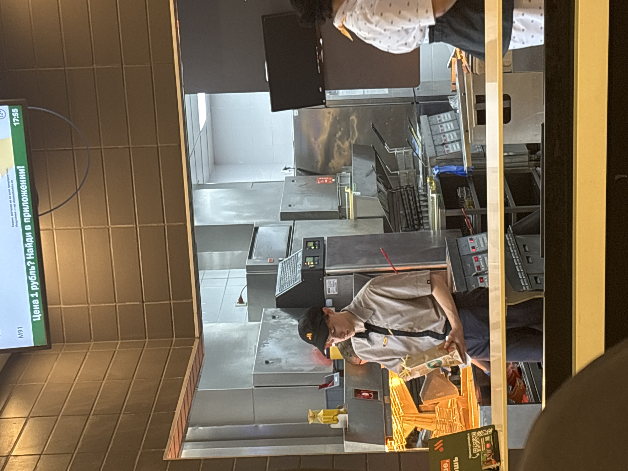 The image shows a McDonald's restaurant interior. A worker wearing a McDonald's uniform and hat is seen preparing food. Another employee wearing a white shirt with black dots is visible in the background. There are several pieces of kitchen equipment and metal surfaces. The location is likely in Vsevolozhsk, Russia, based on the Cyrillic script on the screen in the upper left, which displays an advertisement for McDonald's in Russian. The time on the screen reads 17:55.