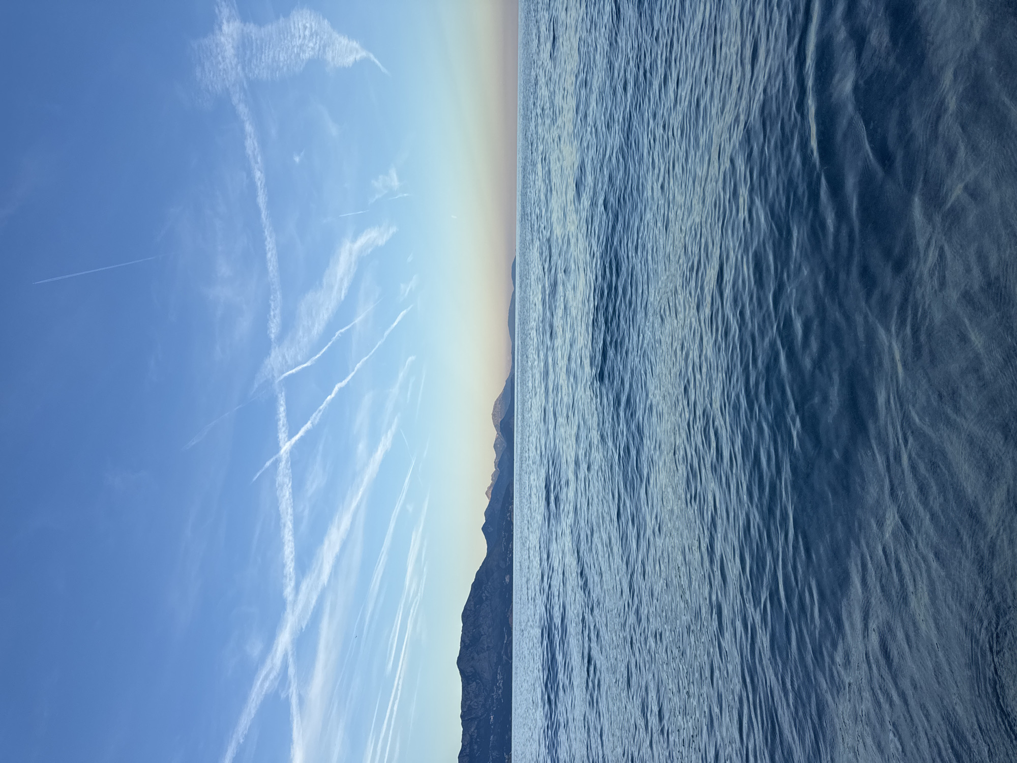 The image depicts a vast coastal seascape under a bright blue sky. The upper two-thirds of the frame show a clear blue sky, transitioning to a lighter, paler blue near the horizon, heavily marked by numerous white aircraft contrails. These contrails vary in density and diffusion, forming several intersecting lines and patterns. A dark, continuous mountain range extends across the middle ground, silhouetted against the lighter sky, with subtle indications of coastal structures or settlements visible on the lower slopes of the mountains to the left. The lower half of the image is dominated by an expanse of deep blue water, displaying gentle, consistent ripples across its surface, which darkens slightly towards the bottom foreground. No individuals, vessels, or specific objects are visible on the water or in the immediate foreground. The scene portrays an open sea with a mountainous coastline, a geographical configuration that does not align with Prijevor, Montenegro, an inland village. Therefore, no specific location within Prijevor can be identified from this image.
