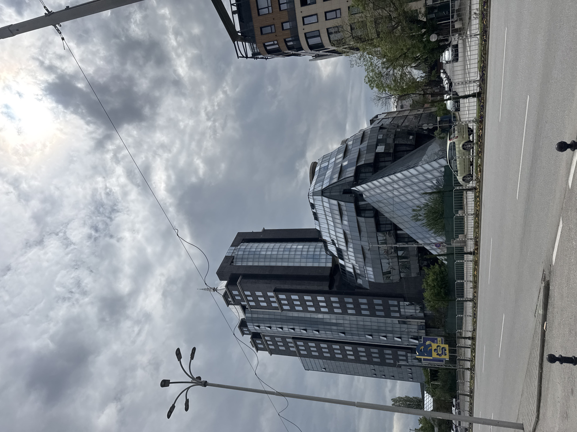 The image shows a street scene in Plovdiv, Bulgaria, featuring the Capital City Center building complex. This complex consists of two modern buildings with geometric designs and glass facades. One building is a high-rise with a rectangular shape, while the other has a more irregular, fragmented form. A street runs alongside the buildings, separated by a metal fence. There are several cars parked along the street. The sky is overcast with gray clouds. Power lines and streetlights are visible.