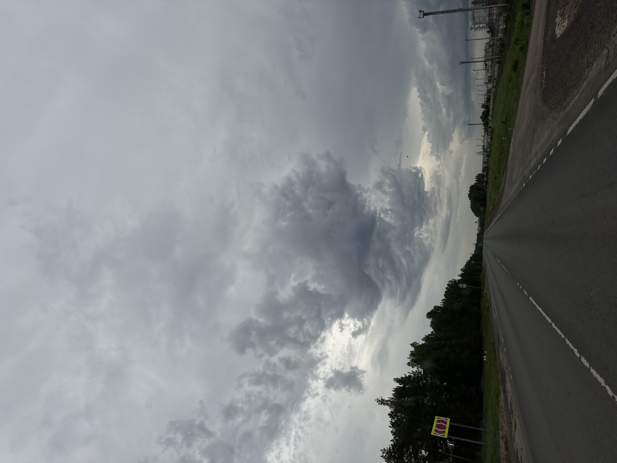 A long asphalt road stretches into the distance under a predominantly overcast sky. The road has a white line marking the edge. To the left of the road is a line of trees and shrubs. There is a yellow and red sign on the roadside. Beyond the trees, industrial structures are visible. The location is Бизяки, Russia.